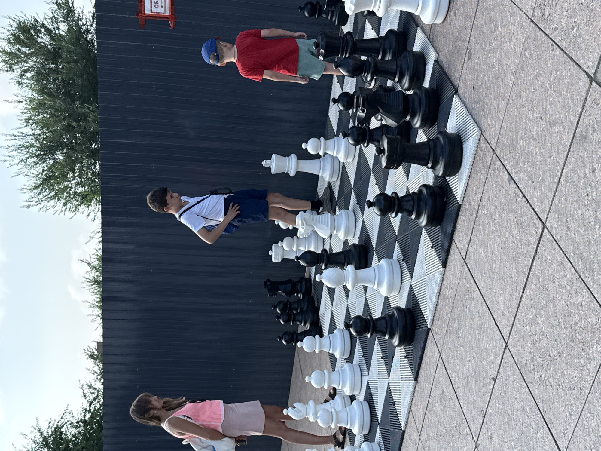 An outdoor, full shot captures three children in casual summer clothing standing near a life-sized chessboard set up on a tiled ground. A young girl on the left, wearing a pink sleeveless top and a light-colored skirt, carries a white backpack. To her right, a boy in a white t-shirt and dark shorts stands with his hands on his hips. Further to the right, a boy in a red t-shirt and light green shorts, wearing a blue baseball cap, faces away from the camera. The giant chess pieces, rendered in black and white, are arranged on a patterned mat. Behind the chess set is a dark, corrugated metal fence, and beyond that, lush green trees and a cloudy sky. The scene is likely set in Vladivostok, Russia, given the context of the image.