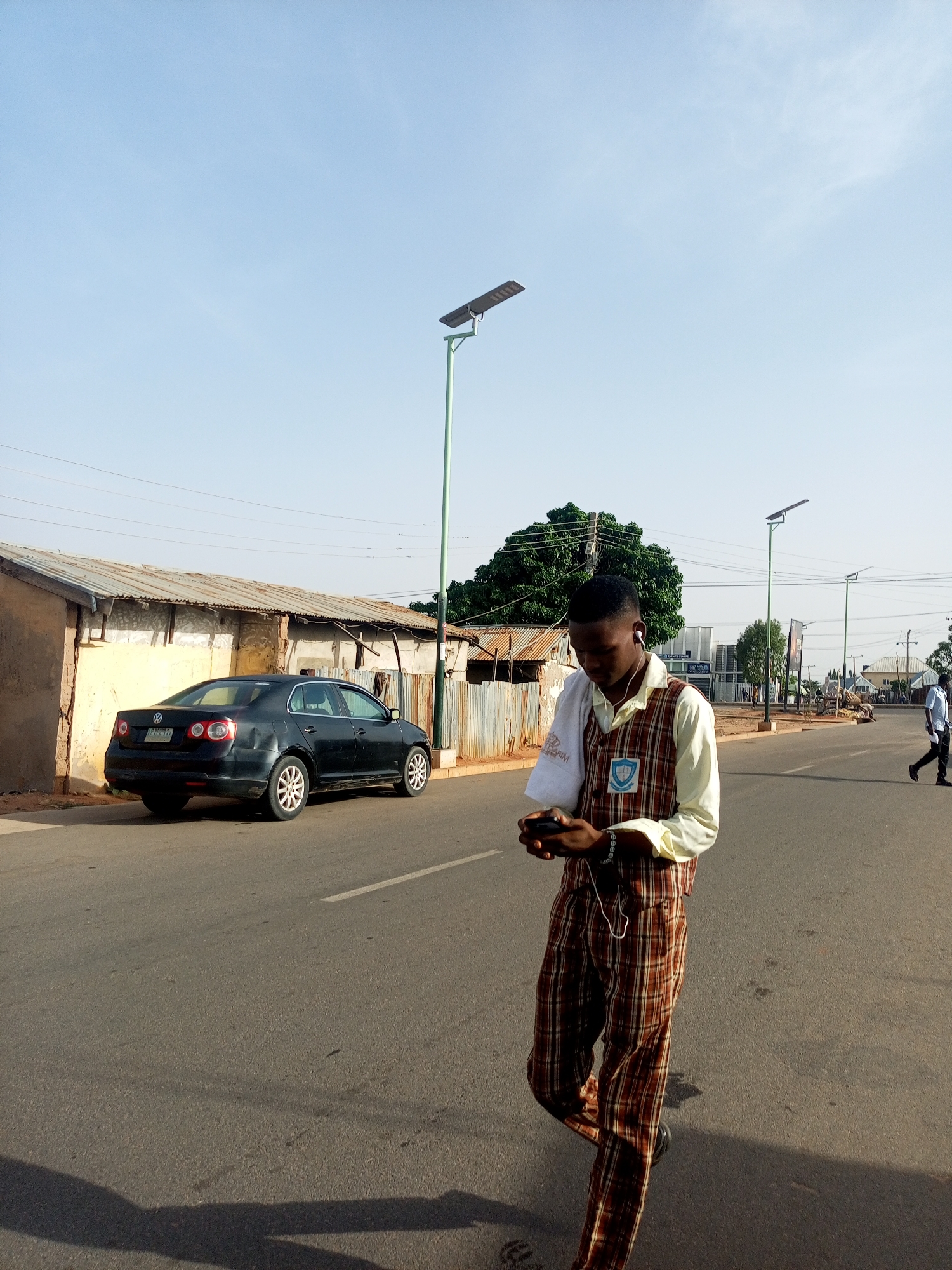 A young man, dressed in a school uniform consisting of a light yellow shirt, a brown and white plaid vest, and matching pants, is walking down a paved road. He is looking at a smartphone in his hands and has earphones connected to the device. To his left, a dark-colored car is parked next to a weathered, one-story building with a corrugated metal roof. Behind the car is a green tree. Further down the road, on the right side, another person is walking away from the camera. Streetlights are placed along the road. The location is Tempure, Nigeria.