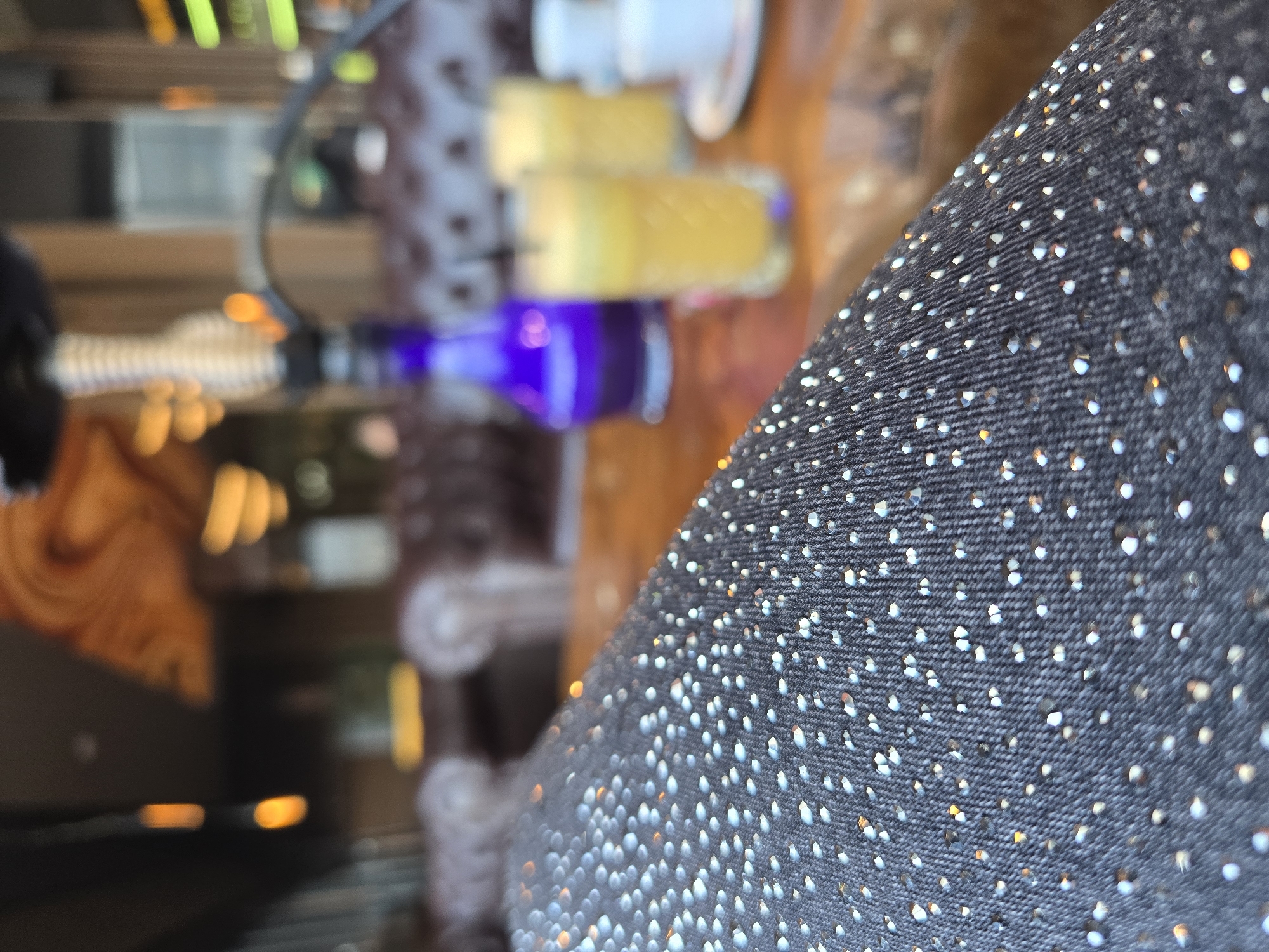 The image shows a close-up of a black fabric with shiny silver-colored dots. In the blurred background, there is a blue bottle, two yellow cylindrical objects on a wooden surface, and a dark object to the left. It is difficult to determine the exact location and any presence of people or actions due to the blur. There is no information provided about the city of Burgas, Bulgaria.
