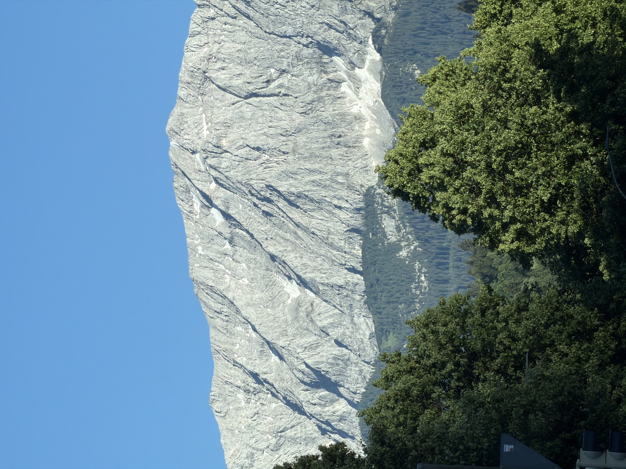 The image shows a majestic, rocky mountain range with snow-capped peaks dominating the background under a clear blue sky. The mountain's surface is textured with numerous deep crevices and slopes, indicative of its rugged natural terrain. In the mid-ground, a dense forest of dark green coniferous trees stretches across the base of the mountains. In the foreground, lush green deciduous trees with full canopies obscure the lower portion of the scene. A traffic signal and part of a dark, angled structure are visible on the far left, suggesting an urban or semi-urban environment bordering the natural landscape. The lighting suggests it is daytime, likely late morning or early afternoon, with the sun casting shadows on the mountain's surface. This scene likely depicts the Zugspitze, Germany's highest peak, as seen from the vicinity of Garmisch-Partenkirchen, a popular Bavarian resort town known for its access to the Alps.