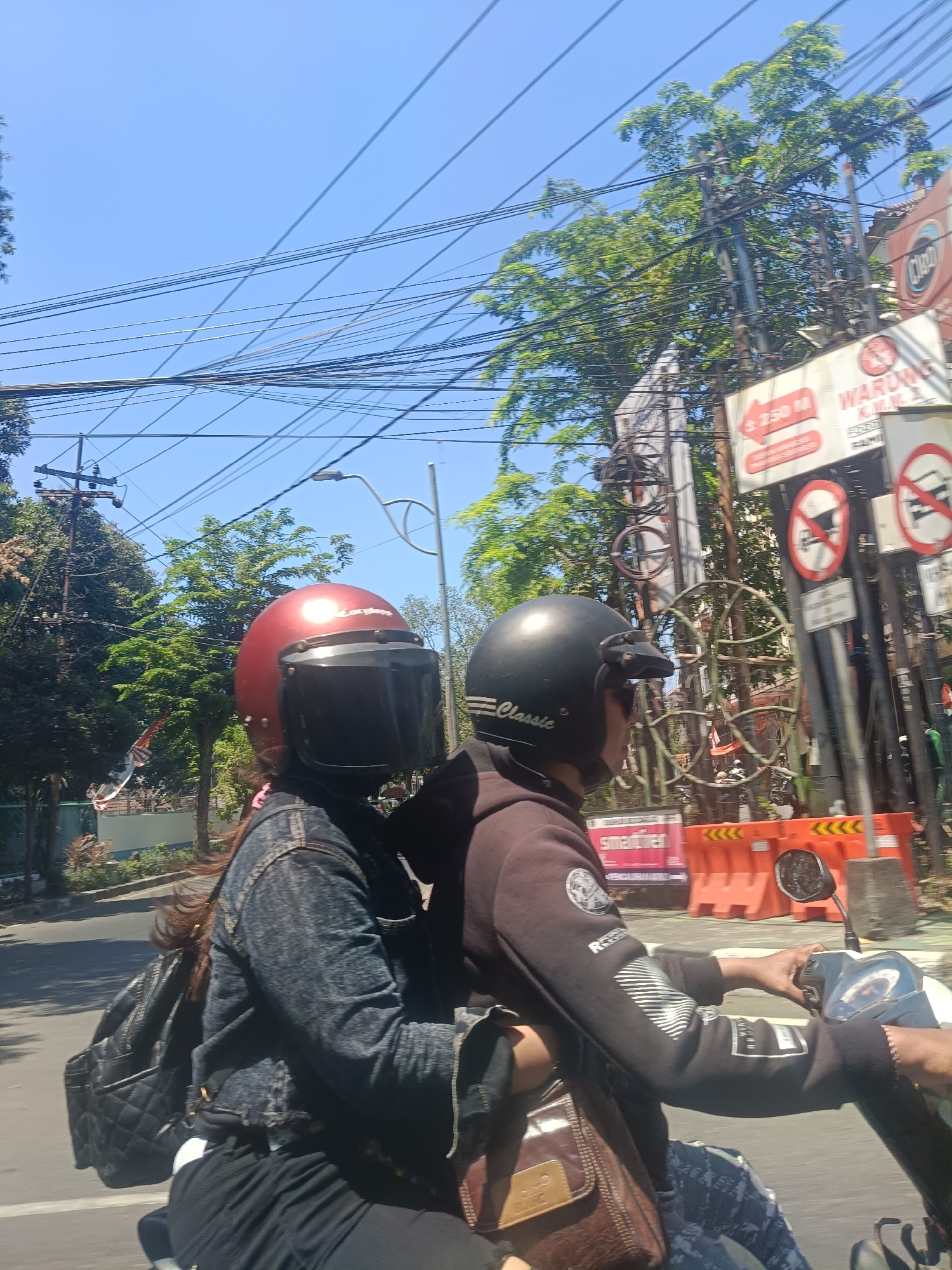 Two individuals are riding a motorcycle on a street in Sidoarjo, Indonesia. The person in front is wearing a black helmet with the brand "Classic" visible and a black hooded sweatshirt. The person behind is wearing a red helmet with a dark visor and a denim jacket. Both are wearing dark pants. The rider in front has their right hand on the handlebar. A brown leather bag is visible on the left side of the motorcycle, and a black backpack is on the back. The environment includes trees, power lines, street signs, and orange traffic barriers. The sky is clear and blue. One sign indicates "WARUNG K.W.M" and another has a distance of "250 M".