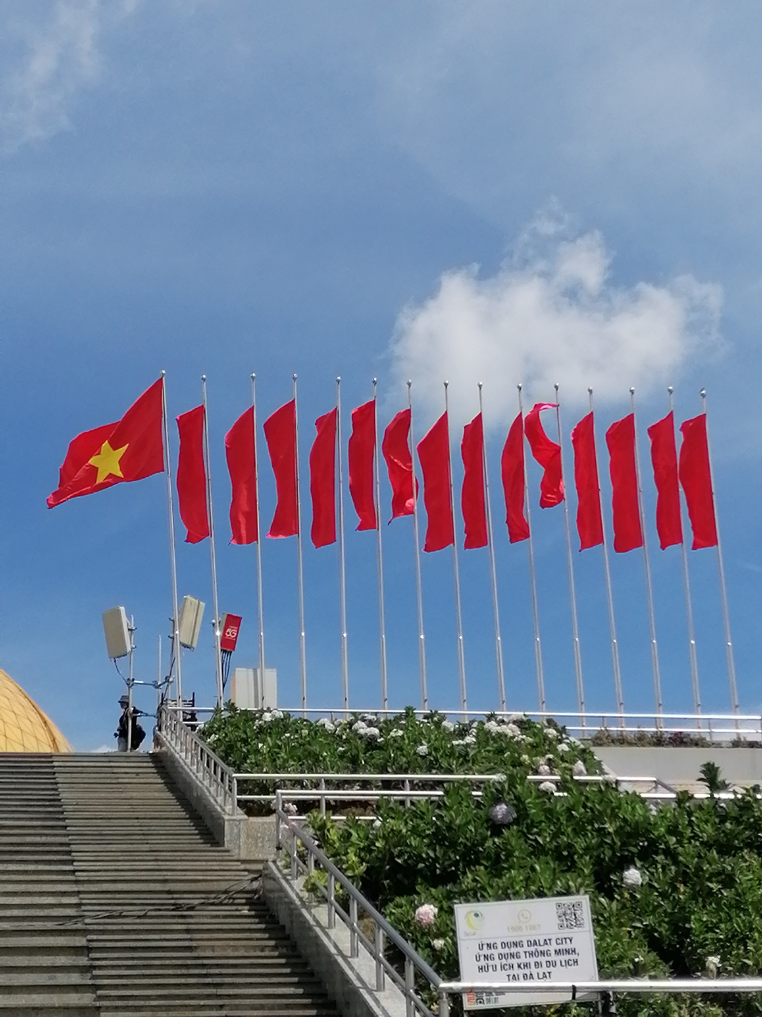 This image captures a bright, sunny day in Da Lat, Vietnam, with a clear blue sky and scattered white clouds. A prominent feature is a line of red flags with a single yellow star on the leftmost flag, fluttering in the breeze. These are likely Vietnamese flags. The flags are mounted on tall, silver poles.
In the foreground, a wide, grey stone staircase leads upwards. To the left of the stairs, a golden dome-shaped structure is partially visible. On the right side of the stairs and behind a metal railing, lush green bushes with white flowers are in bloom, suggesting a well-maintained garden.
Further up and behind the flags, several pieces of electronic equipment, possibly communication towers, are mounted on poles. One has a "5G" logo visible. A person wearing dark clothing and a hat is standing near the top of the stairs, looking towards the flags.
At the base of the green foliage, a white sign with text and a QR code is mounted. The text, which appears to be in Vietnamese, mentions "ỨNG DỤNG DALAT CITY" (Dalat City App) and "ỨNG DỤNG THÔNG MINH, HỮU ÍCH KHI ĐI DU LỊCH TẠI ĐÀ LẠT" (Smart app, useful for traveling in Da Lat).
The overall scene conveys a sense of civic pride and a welcoming atmosphere, possibly at a landmark or public space in Da Lat. The bright sunlight and clear sky indicate it's likely daytime, perhaps late morning or early afternoon.