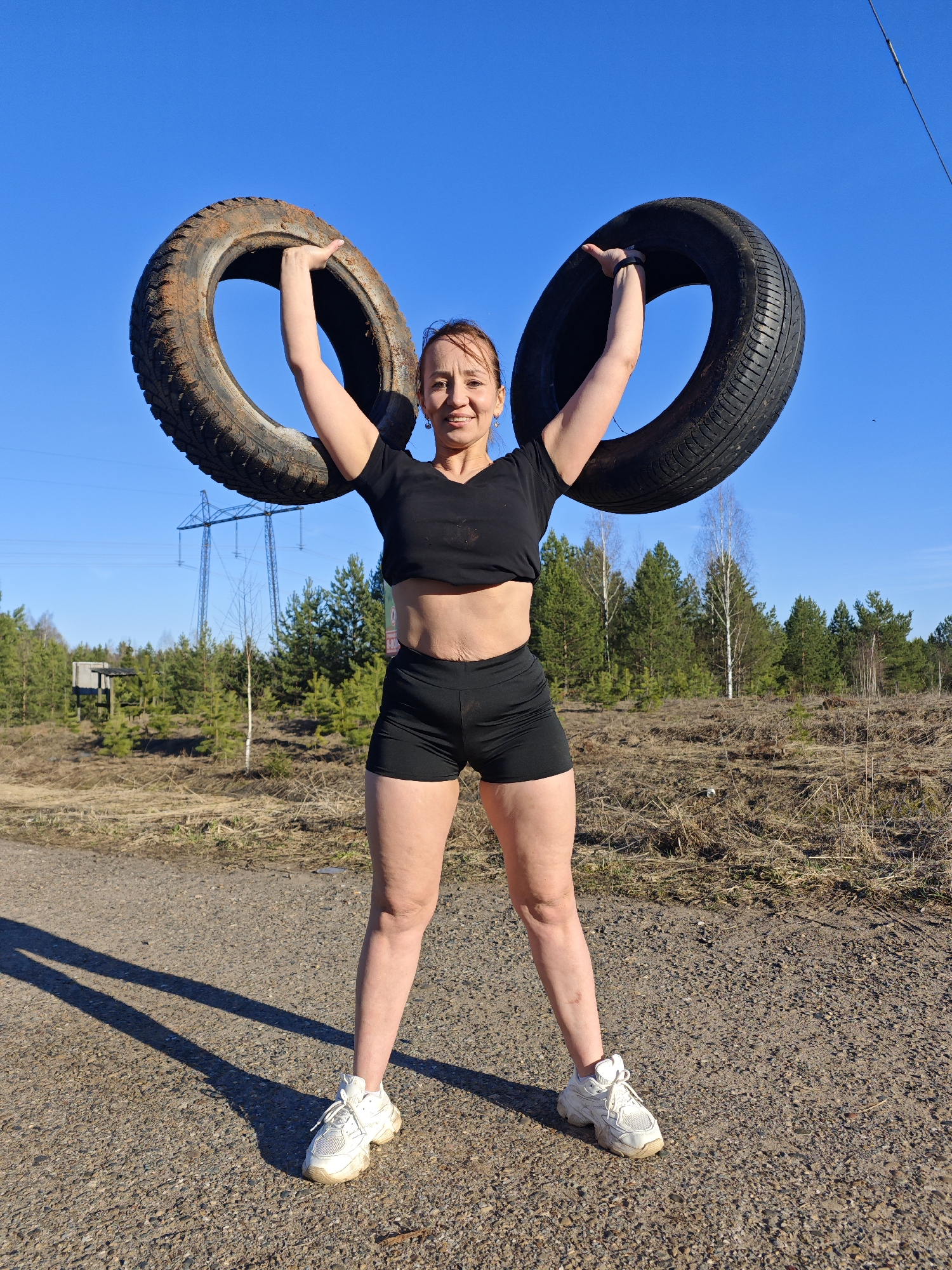 A strong, athletic woman is captured mid-action, holding two vehicle tires above her head against a clear, bright blue sky. She stands on a dirt and gravel road, directly facing the camera with a determined yet cheerful smile. She is wearing a black short-sleeved crop top that exposes her midriff and black athletic shorts, paired with white sneakers. Her arms are flexed as she supports the heavy tires; the left tire appears to be a dirty, possibly winter tire with a rugged tread, while the right tire seems a bit cleaner with a summer tread.
The setting is an open, rural or semi-rural landscape. In the background, there is sparse woodland consisting of pine and birch trees, with dry, light-colored grass and bushes covering the ground. Prominently visible in the mid-distance to the left, a large metal electricity pylon stands tall against the sky, indicating an industrial or utility presence in the natural environment. The bright sunlight casts long shadows on the road from the woman and the tires, suggesting it is either morning or late afternoon on a clear day. No discernible text is visible on the tires, clothing, or in the background elements. The scene conveys a sense of outdoor strength training, likely in the Chaikovsky region of Russia.