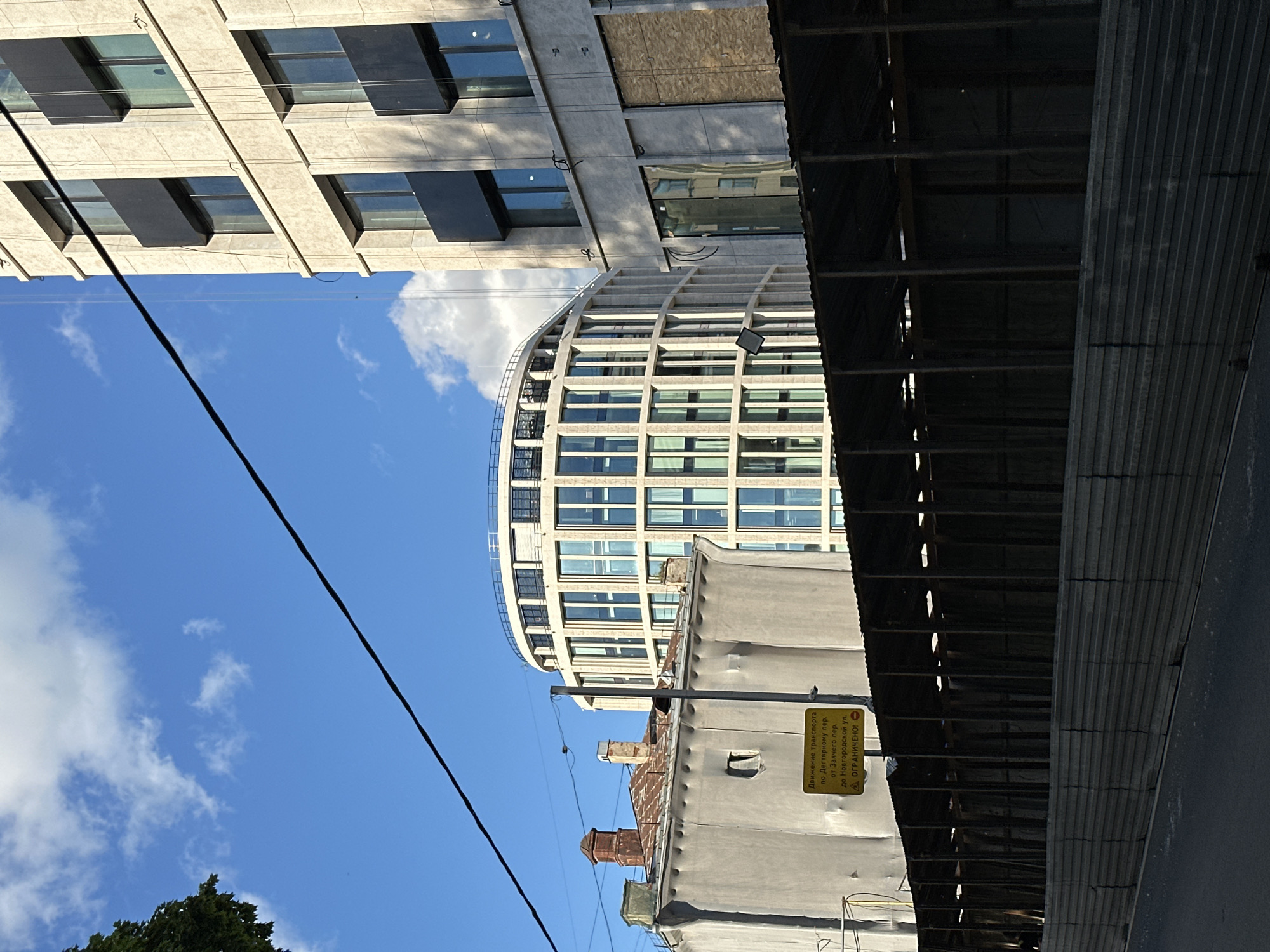 This is a photograph taken at street level in Saint Petersburg, Russia, showing an urban scene dominated by modern architecture and construction. In the foreground, a temporary, dark metal fence topped with a corrugated metal roof structure suggests ongoing construction or a street market. Behind this, two distinct buildings are visible. To the left, an older, gray building with a weathered, rusty metal roof and a small, recessed window stands. A yellow sign with Cyrillic text and an exclamation mark warning symbol is mounted on a pole in front of this building. To the right of the older building and slightly further back is a strikingly modern, cylindrical building with a facade of numerous vertical white fins framing large windows. Further to the right, a taller, rectangular building with a lighter stone facade and dark, recessed window sections rises. The sky above is blue with scattered white clouds, and a black electrical wire bisects the upper portion of the frame. The scene is captured on a clear day.