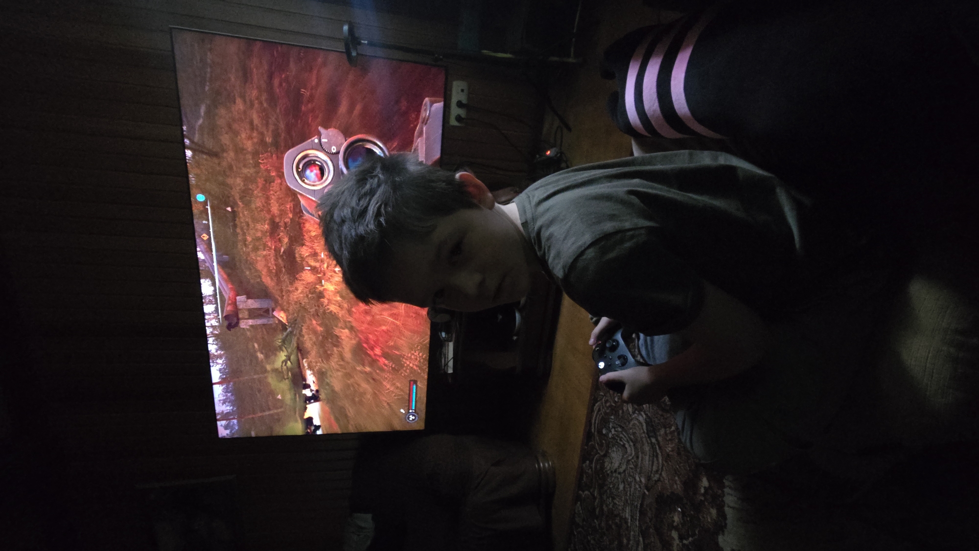 A young boy is playing a video game in a dimly lit room. He is positioned to the right of a large screen displaying the game. The game appears to be a first-person shooter, with a scope from a weapon visible in the foreground of the screen. The boy is holding a video game controller in his right hand. He is wearing a dark t-shirt. Behind him, a person is partially visible, wearing a black shirt with pink stripes. The setting is indoors, with wooden paneling on the wall to the left of the screen. The overall mood is dark and focused on the game. The time of day is indeterminate due to the artificial lighting and dark atmosphere. There is no visible text in the image.