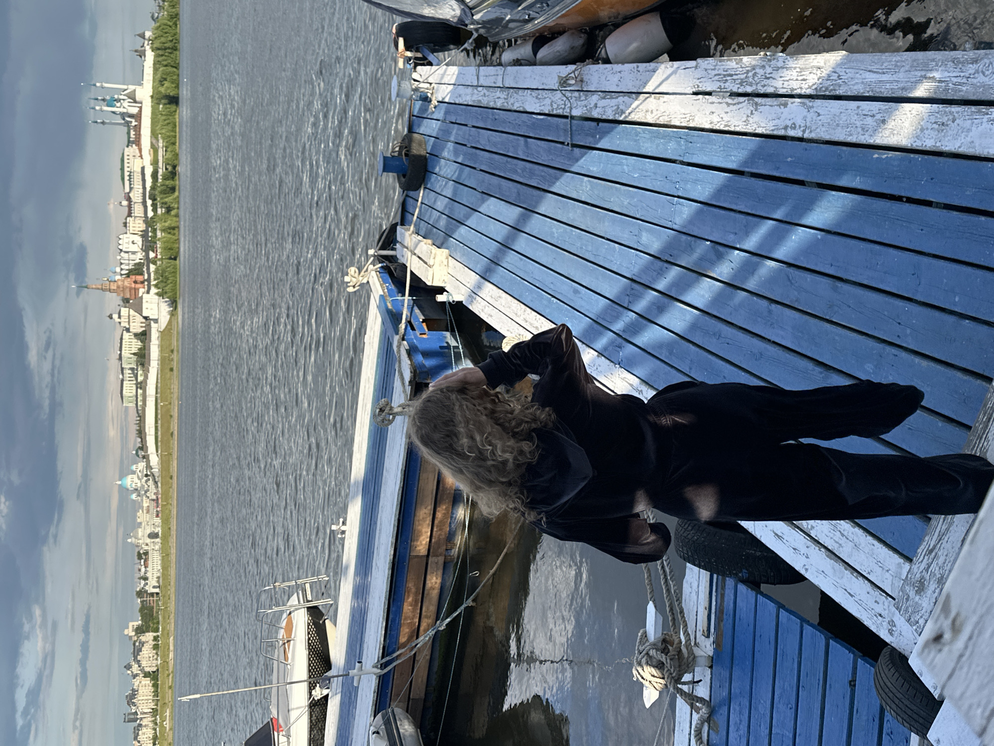 A woman with blonde curly hair, dressed in black, stands on a blue and white painted wooden dock, looking out at a body of water. On the opposite shore is the city of Kazan, Russia, recognizable by its distinctive skyline, including the white-walled Kremlin with its blue-domed churches and minarets. Several boats and tires are visible around the dock.