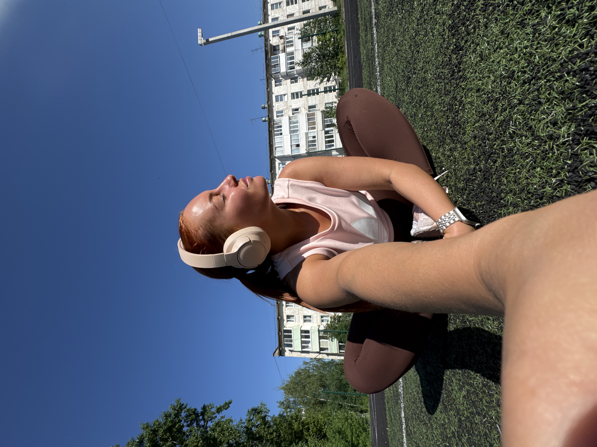 A fair-skinned person with long red hair sits on a grass-like surface under a bright blue sky in what appears to be a selfie. They wear headphones, a light pink tank top, and brown leggings. A metal watch is visible on their wrist.
The background includes a multi-story building and a light pole, suggesting an urban setting. Based on the architecture, the location is likely in Chaikovsky, Russia.
