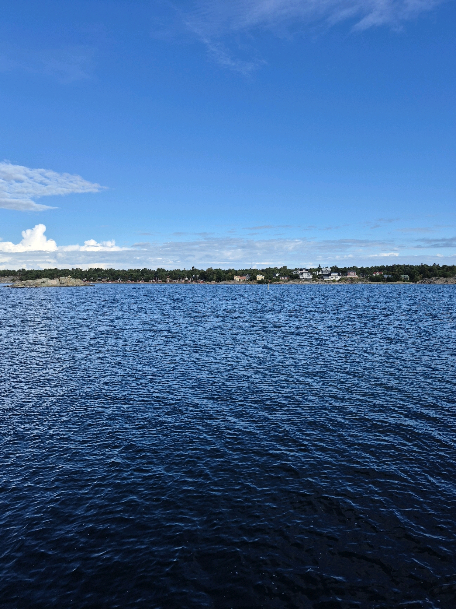 The image depicts a scenic view of the sea in Hanko, Finland. The foreground is dominated by the deep blue, rippling surface of the water, reflecting the bright sky above. In the middle ground, a rocky shoreline is visible, lined with dense green trees. Beyond the trees, a cluster of buildings, likely houses and possibly some red cottages, forms a settlement along the coast. The sky is a vibrant blue with scattered white clouds, adding to the serene atmosphere of a clear day. The image captures a coastal landscape characteristic of the Hanko region, known for its archipelago and maritime charm.