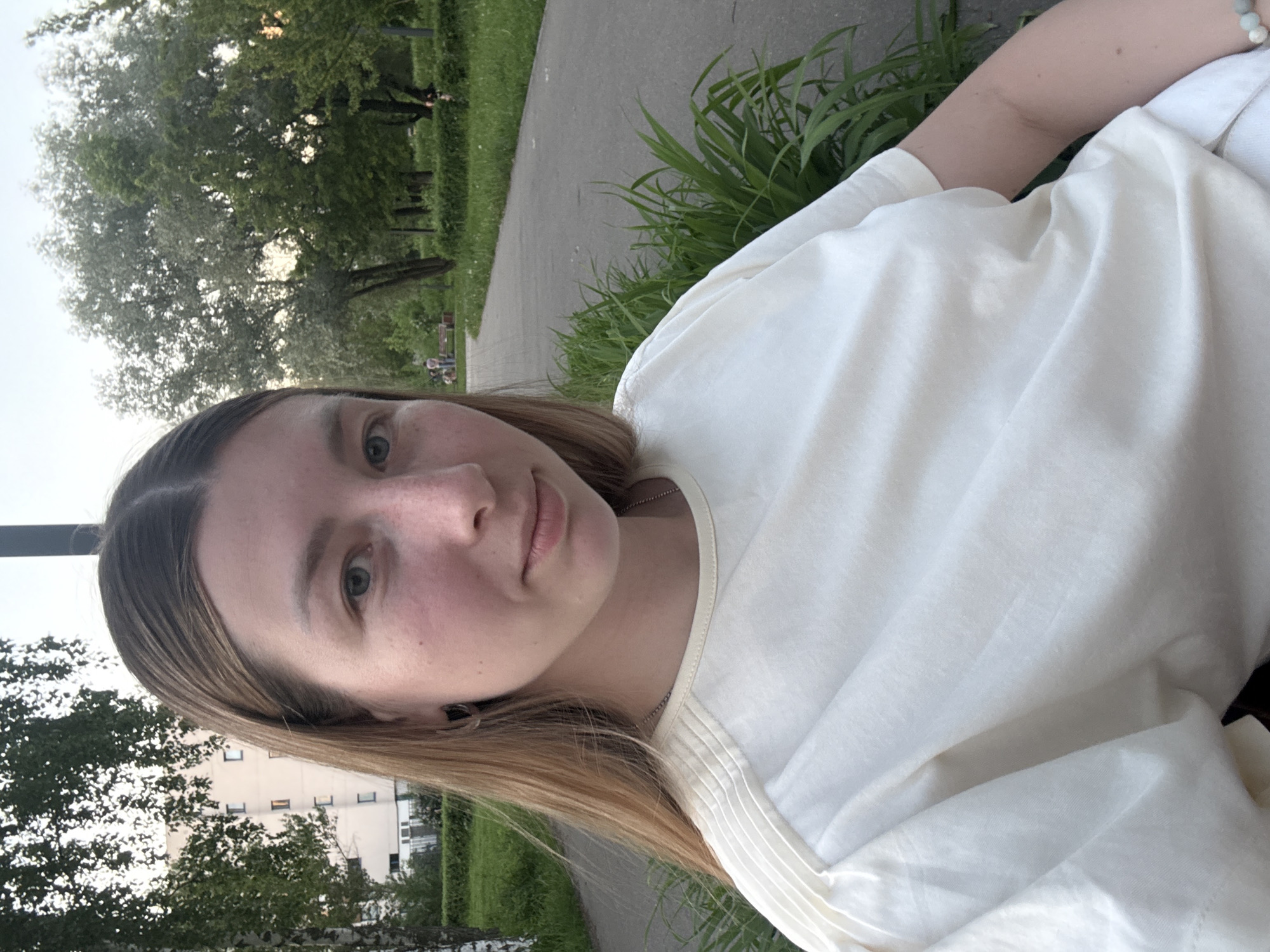 The image shows a person with light brown hair and fair skin wearing a white shirt. The person is outdoors, with trees and a paved path visible in the background. A building is partially visible through the trees. The scene appears to be in a park or green space. Without further information, pinpointing a specific location in Moscow is impossible.