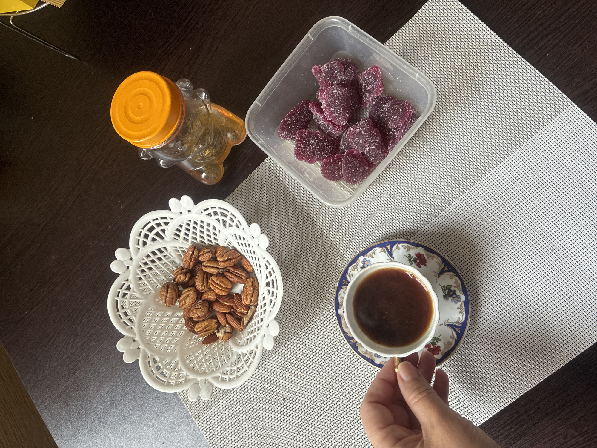 A tabletop still life features a cup of coffee, a container of dark purple sugar-coated candies, a white lacy bowl of pecans and almonds, and a honey bear-shaped jar with an orange lid. The objects are arranged on a textured white place mat, set against a dark brown tabletop. A hand reaches for the coffee cup. The location may be Sochi, Russia, based on the image context.