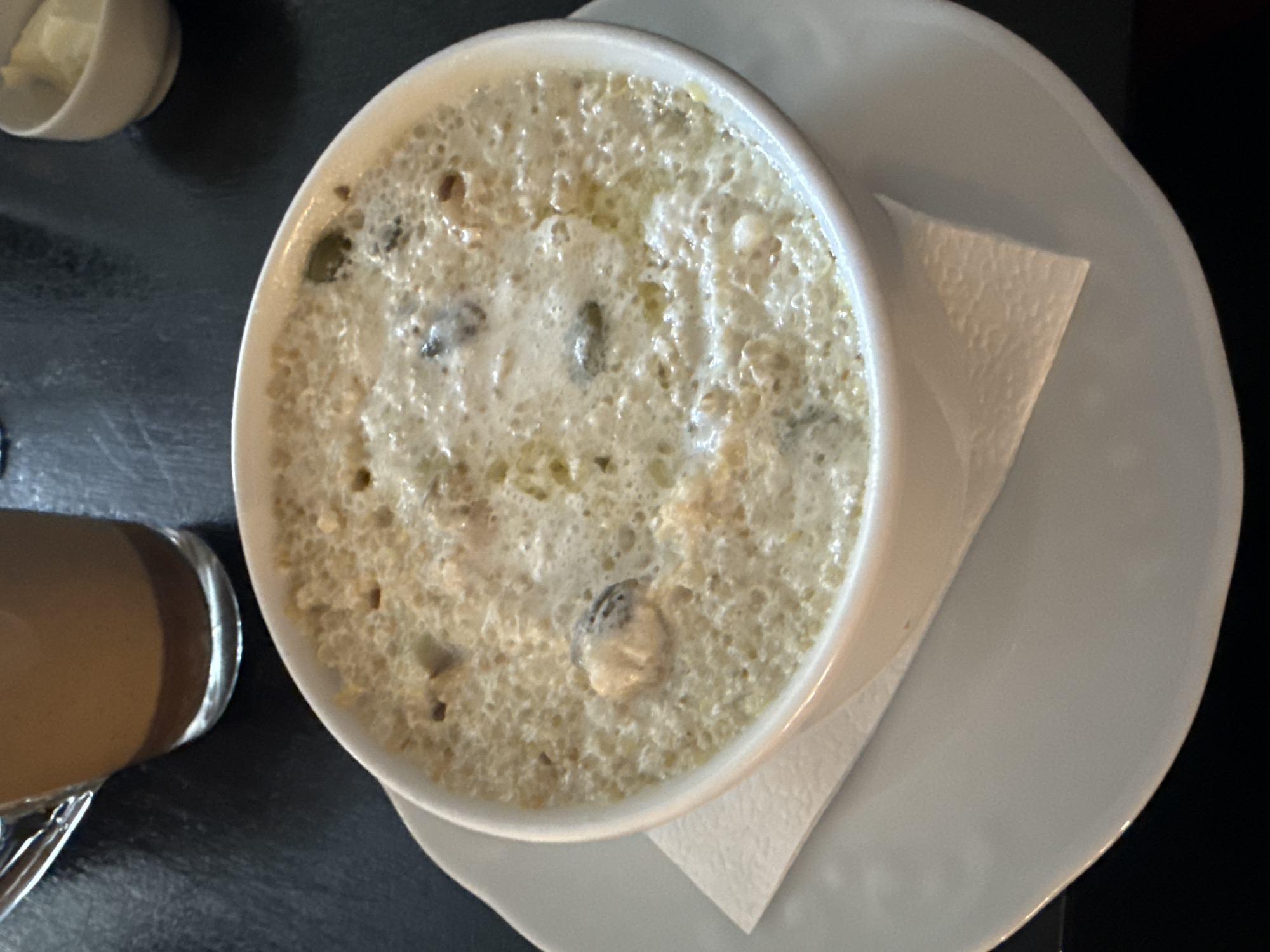 The image shows a bowl of oyster stew served on a white plate with a napkin beneath it. A cup with a light-colored liquid is visible to the left, and a small bowl is visible in the upper left corner. The background is a dark surface. The photo was taken in Ust-Koksa, Russia.