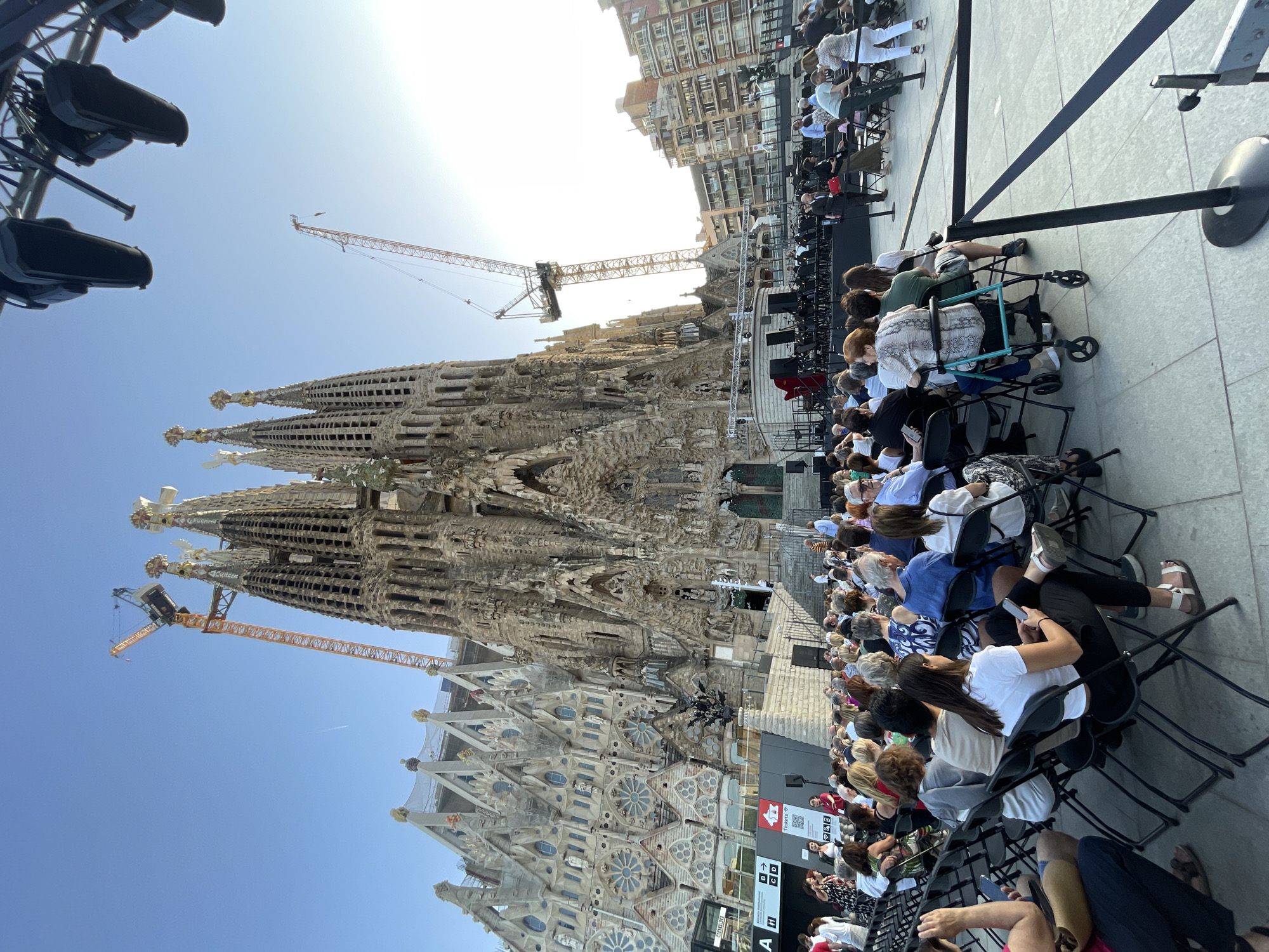 The image depicts an outdoor event in front of the Sagrada Familia in Barcelona, Spain. A large audience is seated on chairs, facing a stage set up with musical instruments and what appears to be an orchestra. The iconic Sagrada Familia, a basilica designed by Antoni Gaudí, is visible in the background, with construction cranes indicating ongoing work. The sky is clear and blue, suggesting a daytime event. People are dressed casually, and some are holding cell phones. The atmosphere appears to be one of a cultural performance or concert.