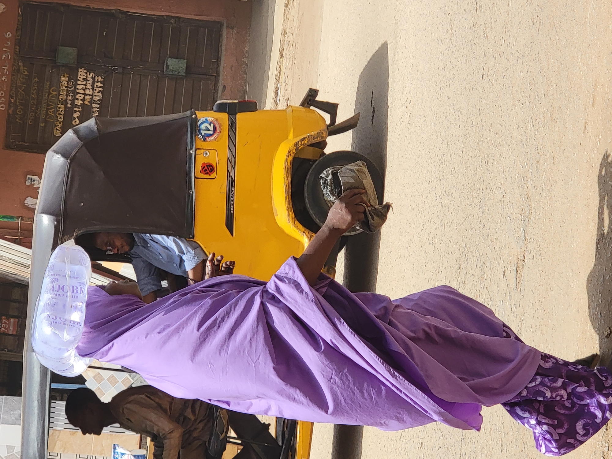 The image shows a man in Maiburiji, Nigeria, cleaning the tire of a yellow and black auto-rickshaw labeled "DELUXE". The man is wearing a large purple robe and a white head covering. Another man is seated in the driver's seat of the auto-rickshaw. The background includes a brown shuttered door with handwritten text and numbers and a tiled wall. The image appears to be taken outdoors on a sunny day.