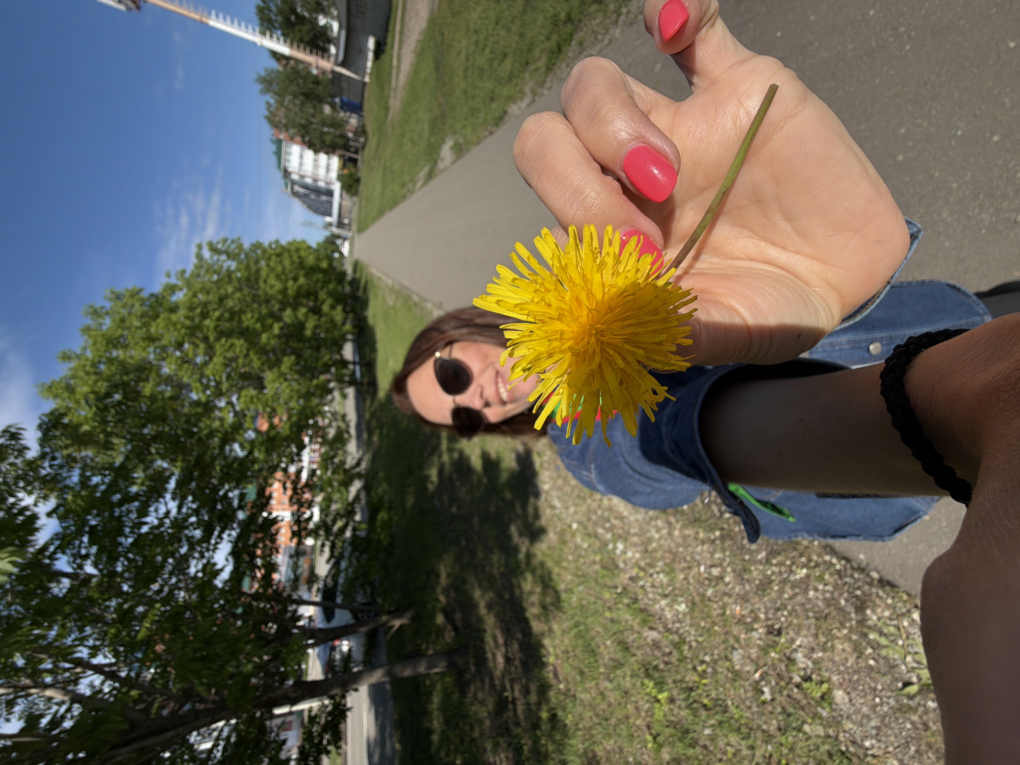 A woman is holding a yellow dandelion in her outstretched hand, the flower filling the foreground. She is wearing sunglasses and a denim jacket, and her fingernails are painted bright pink. The background shows a paved walkway lined with green grass and trees, with buildings and a bright blue sky visible in the distance. The overall impression is a sunny day outdoors.