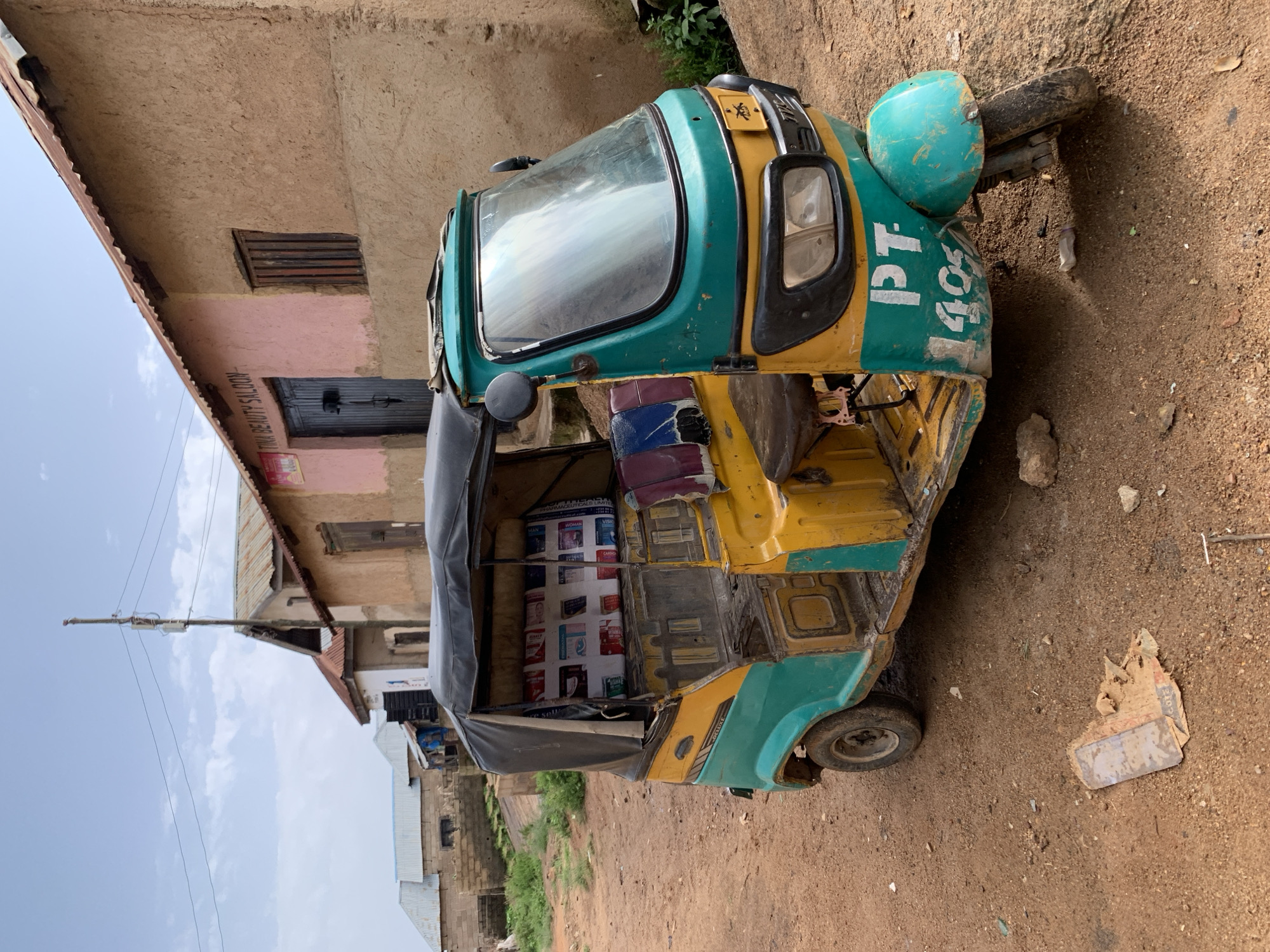 In Jos, Nigeria, a weathered yellow and teal auto-rickshaw is parked on a dirt road in front of a pink and tan building. The building has a corrugated metal roof and a sign that reads "TINA BEAUTY SALOON". The auto-rickshaw has a black canvas canopy and is adorned with various advertisements on its interior backrest. The vehicle's front fender displays the markings "PT 1405" in white lettering. The ground is covered in reddish-brown dirt, with scattered debris, including a torn piece of paper or cardboard. In the background, other buildings with metal roofs are visible, and a tall utility pole stands against a partly cloudy sky. The scene appears to depict a typical street view in a residential or commercial area of Jos.