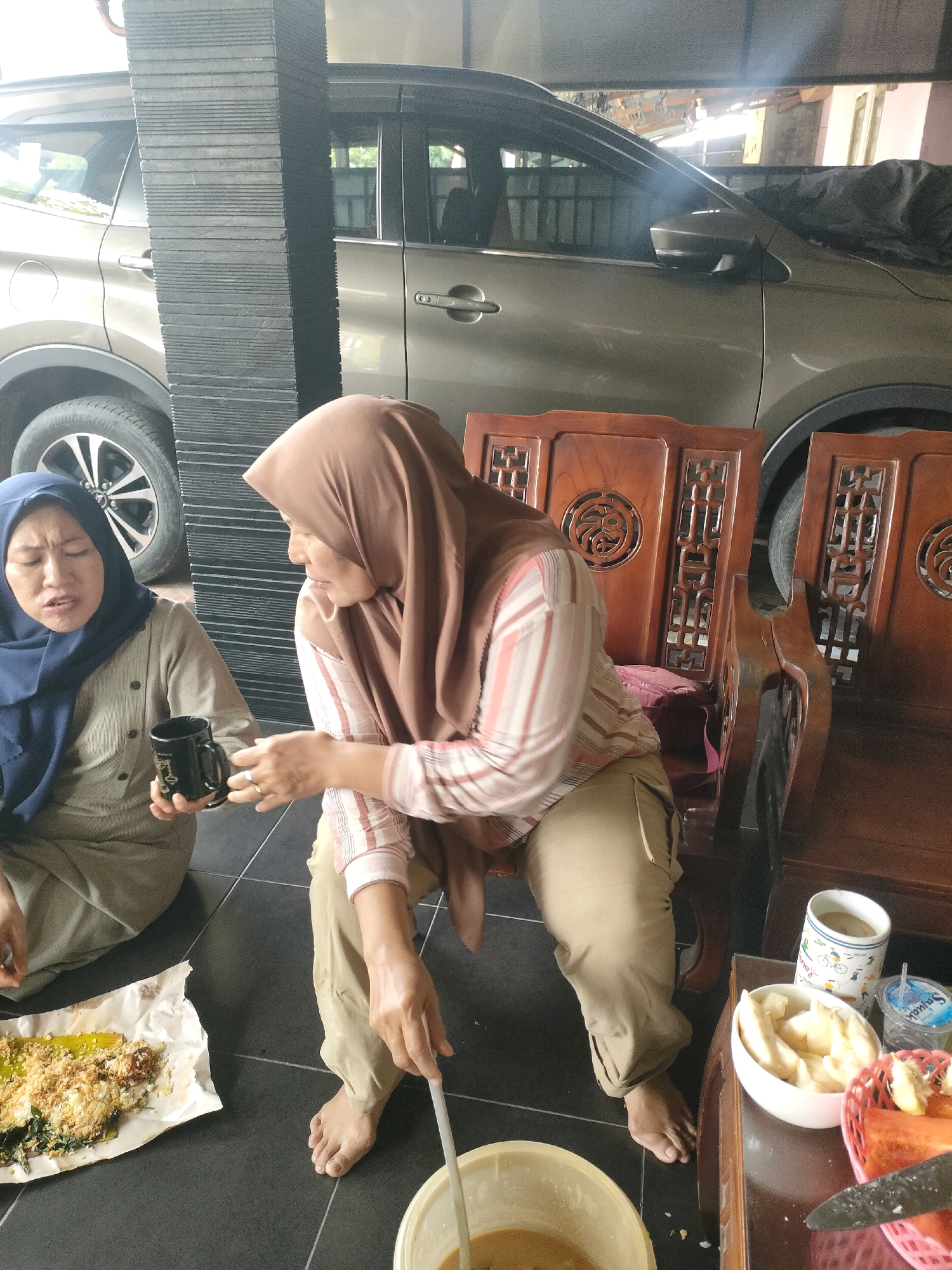 The image depicts an outdoor scene in Pekanbaru, Indonesia. Two individuals are present. The person on the left, wearing a blue hijab and a light brown shirt, is seated on the floor, holding a black mug. The person on the right, wearing a brown hijab and a striped long-sleeved shirt, is seated on a wooden chair and is reaching towards a table. The table holds several items, including a white bowl with sliced fruit, a small cup, and a bottle of water. In the foreground, there is a plate of food on a piece of white paper. A large pillar, constructed from dark, textured blocks, is visible on the left side of the image, partially obscuring a light-colored car parked in the background. The ground appears to be tiled with dark squares. The overall atmosphere suggests a casual gathering or meal.