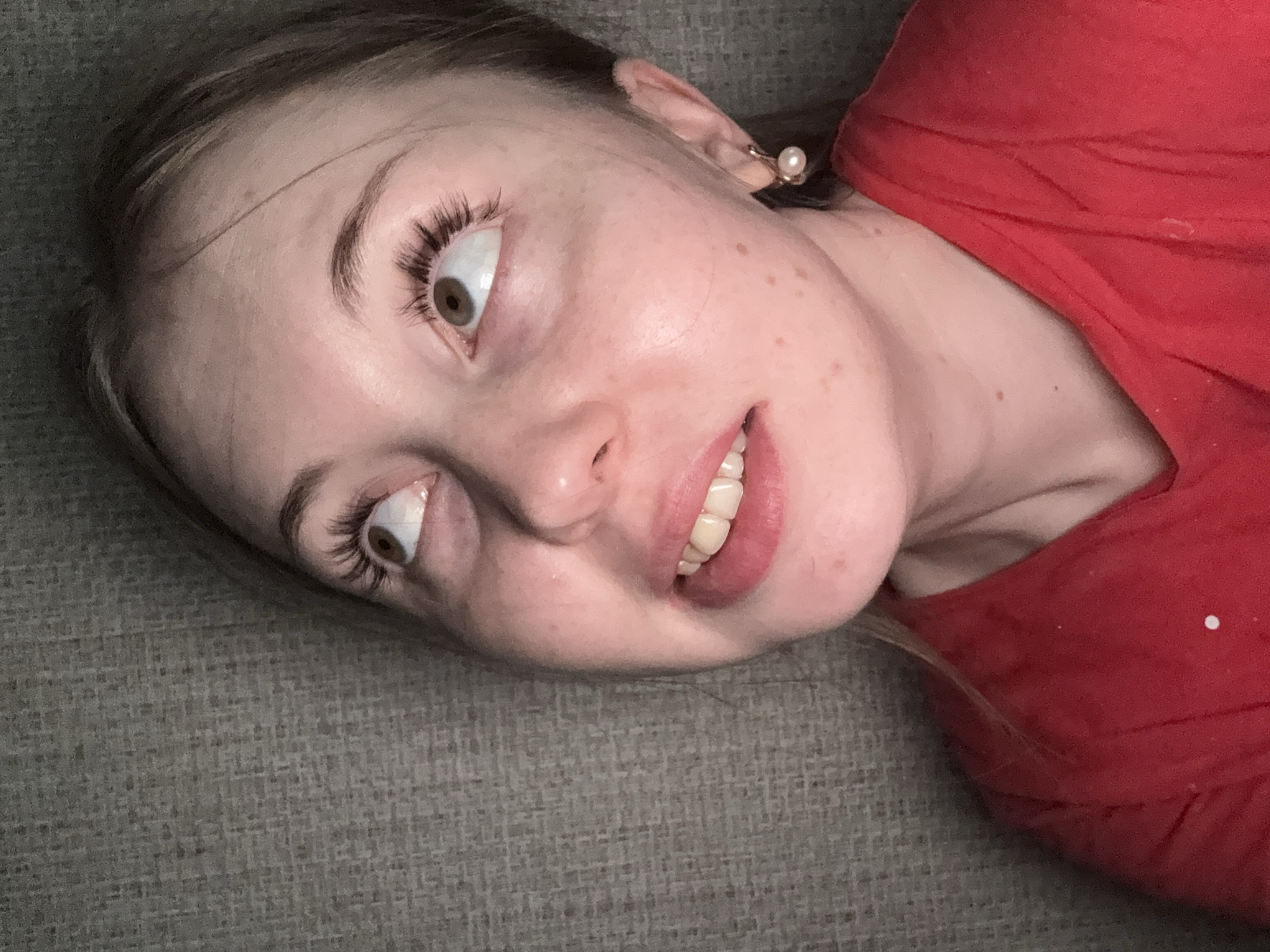 A close-up, angled portrait captures a young woman lying on a textured, grey-green surface, likely a couch or blanket. Her face dominates the frame, positioned diagonally. Her eyes are rolled significantly upwards, revealing the whites and only a small portion of her brown irises, adorned with long, dark eyelashes. Her eyebrows are well-defined. A slight smile plays on her lips, showing her upper teeth. Her skin features light freckles scattered across her cheeks and forehead. She wears a red garment, visible on her right side, and a small, spherical pearl earring is prominent on her right earlobe. The expression on her face, with the upward-rolled eyes and gentle smile, suggests a playful, humorous, or perhaps exaggeratedly exasperated mood. No text is visible in the image.