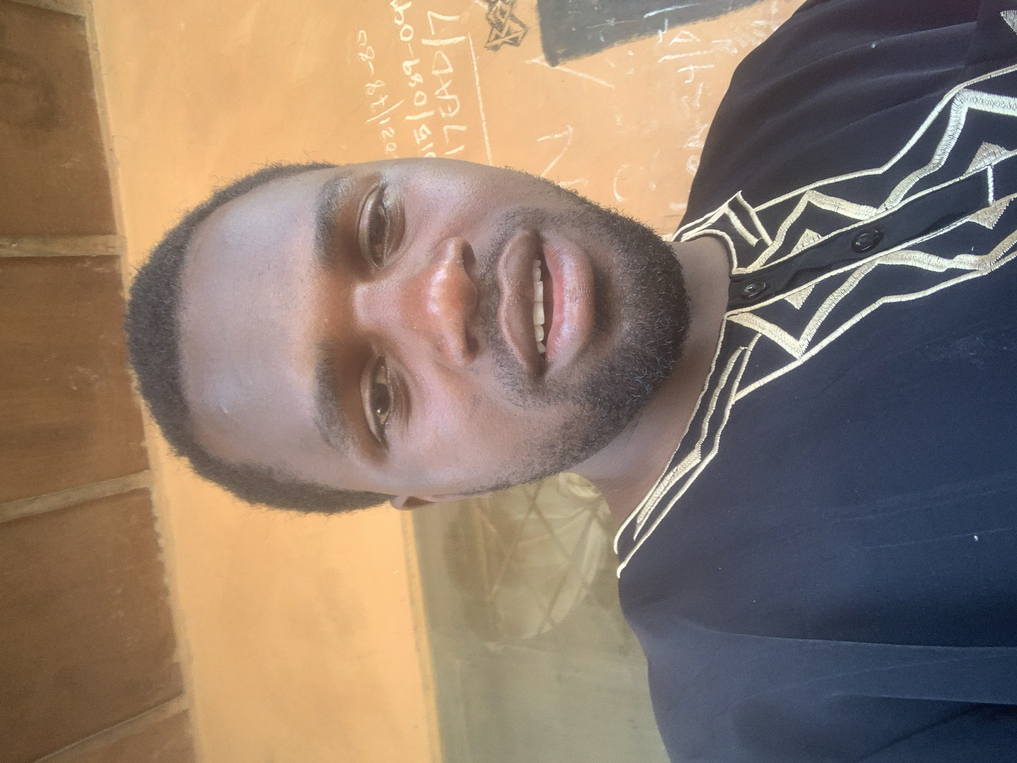 The image shows a close-up selfie of a Black man in his late 20s or early 30s. He has dark skin, short black hair, and a beard. He is wearing a black traditional garment with gold embroidery around the neckline and down the front. The garment appears to be a dashiki or a similar style of West African clothing. The man is looking directly at the camera with a neutral expression, his mouth slightly open.
The background is an orange-colored wall with some writing and symbols scrawled on it in white. The writing appears to be in chalk or paint. One visible line of text reads "021/78-80". There are also some geometric shapes and what looks like a star symbol. The ceiling, visible at the top of the image, is made of wood planks.
Based on the style of clothing and the visible writing, the image was likely taken in Nigeria. The context of the writings on the wall suggests a local setting, possibly a home or a public building. However, without more specific identifying features or context, it is not possible to pinpoint the exact location within Jos, Nigeria.