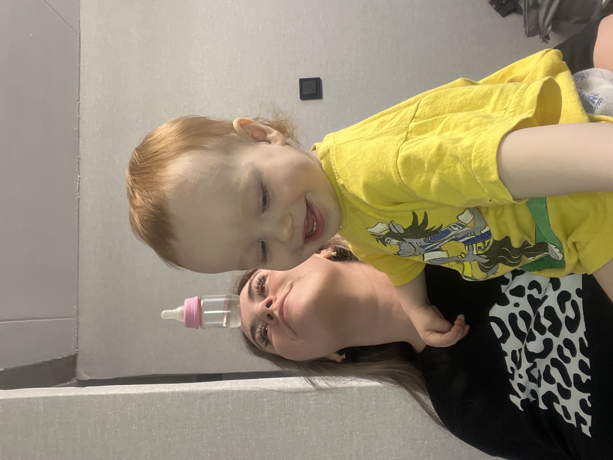 A baby in a yellow cartoon t-shirt lies on a woman wearing a black t-shirt with a cow print design, while holding a baby bottle. The setting appears to be indoors with light gray walls, including a light switch. The location is the city of Chaikovsky, Russia.