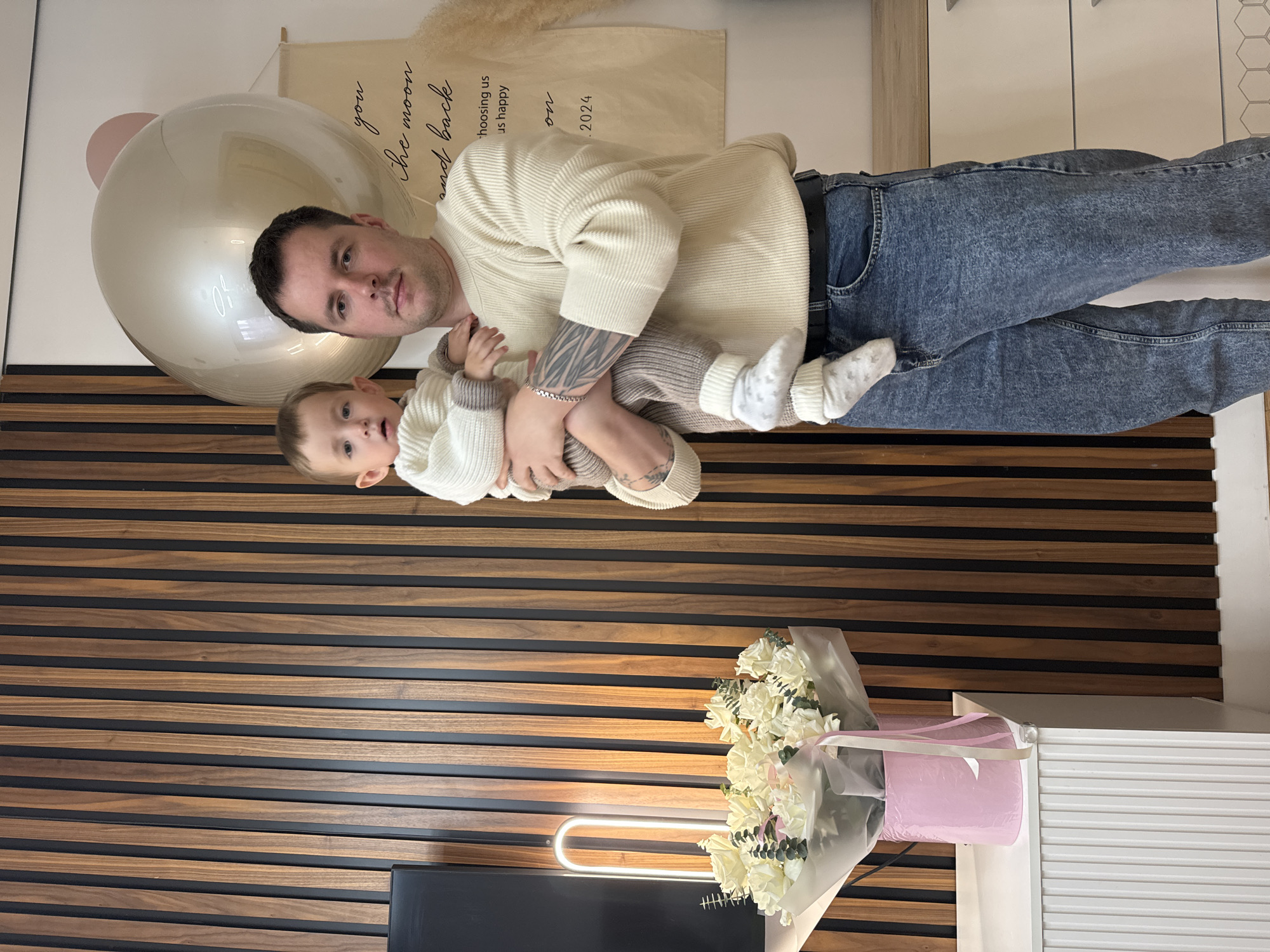 The image shows a man holding a baby indoors. The man is wearing a cream-colored sweater and blue jeans. He has tattoos on his left arm. The baby is wearing a matching cream-colored sweater and pants with socks. Behind the man is a large, translucent balloon and a fabric banner with text. In the foreground, there is a bouquet of white flowers in a pink vase, sitting on a white cabinet, in front of a wall with horizontal wood paneling. A light fixture is visible beneath the bouquet. The image was likely taken in Akulovo (Gp Kubinka), Russia.