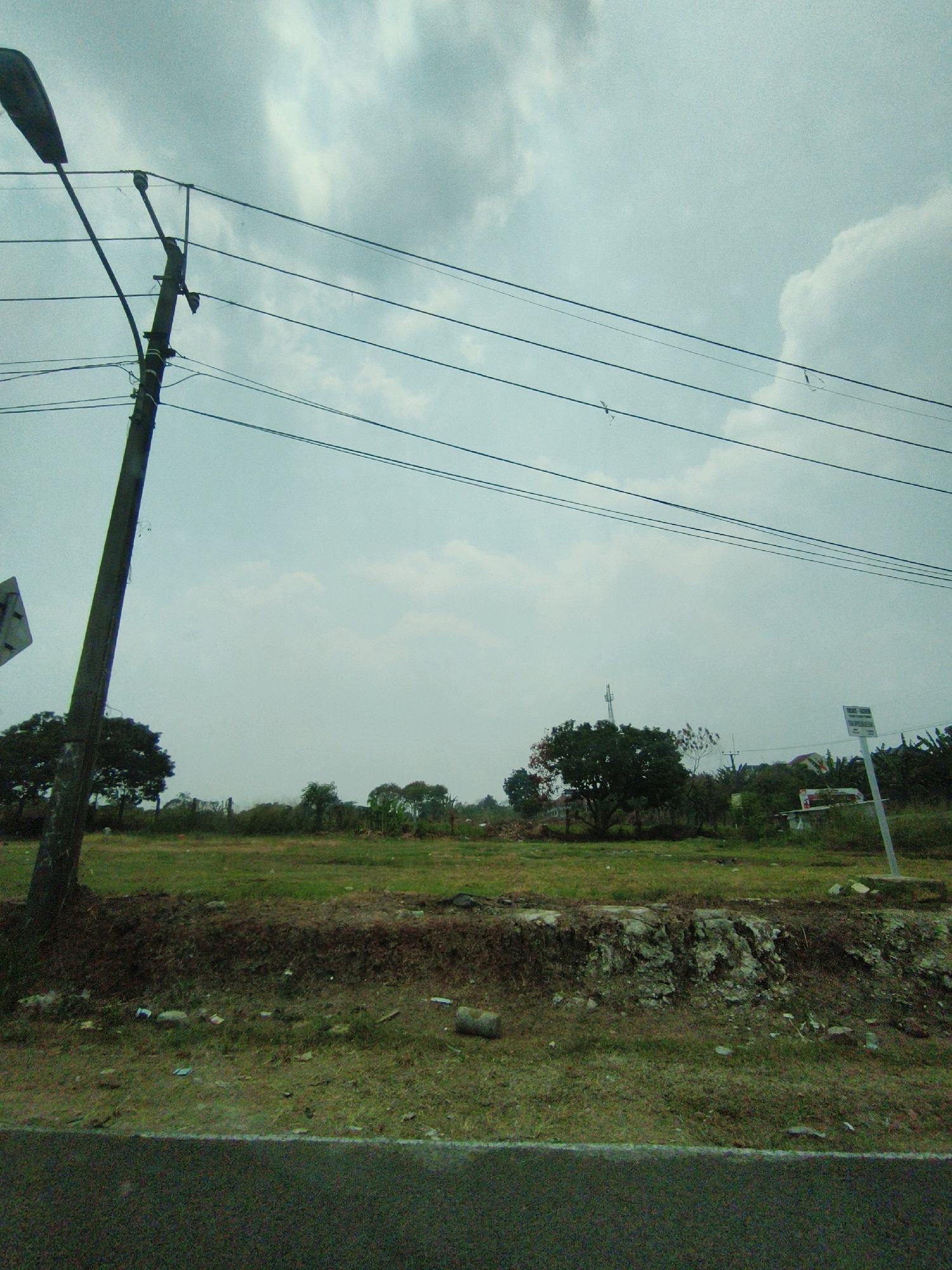 A street-level, eye-level, medium shot captures a desolate, overcast day in Sukaraja, Indonesia. The foreground shows a paved road, with a narrow, grassy shoulder littered with debris. Beyond the shoulder, a dry, unkempt embankment is visible, also strewn with trash. The midground is a grassy field, bordered by a row of trees and vegetation. In the distance, a cell tower stands tall amongst the trees, with a few structures and more trees dotting the landscape. A street light pole with multiple wires extends diagonally across the frame, partially obscuring the cloudy, pale blue sky. The scene conveys a sense of neglect and underdevelopment.