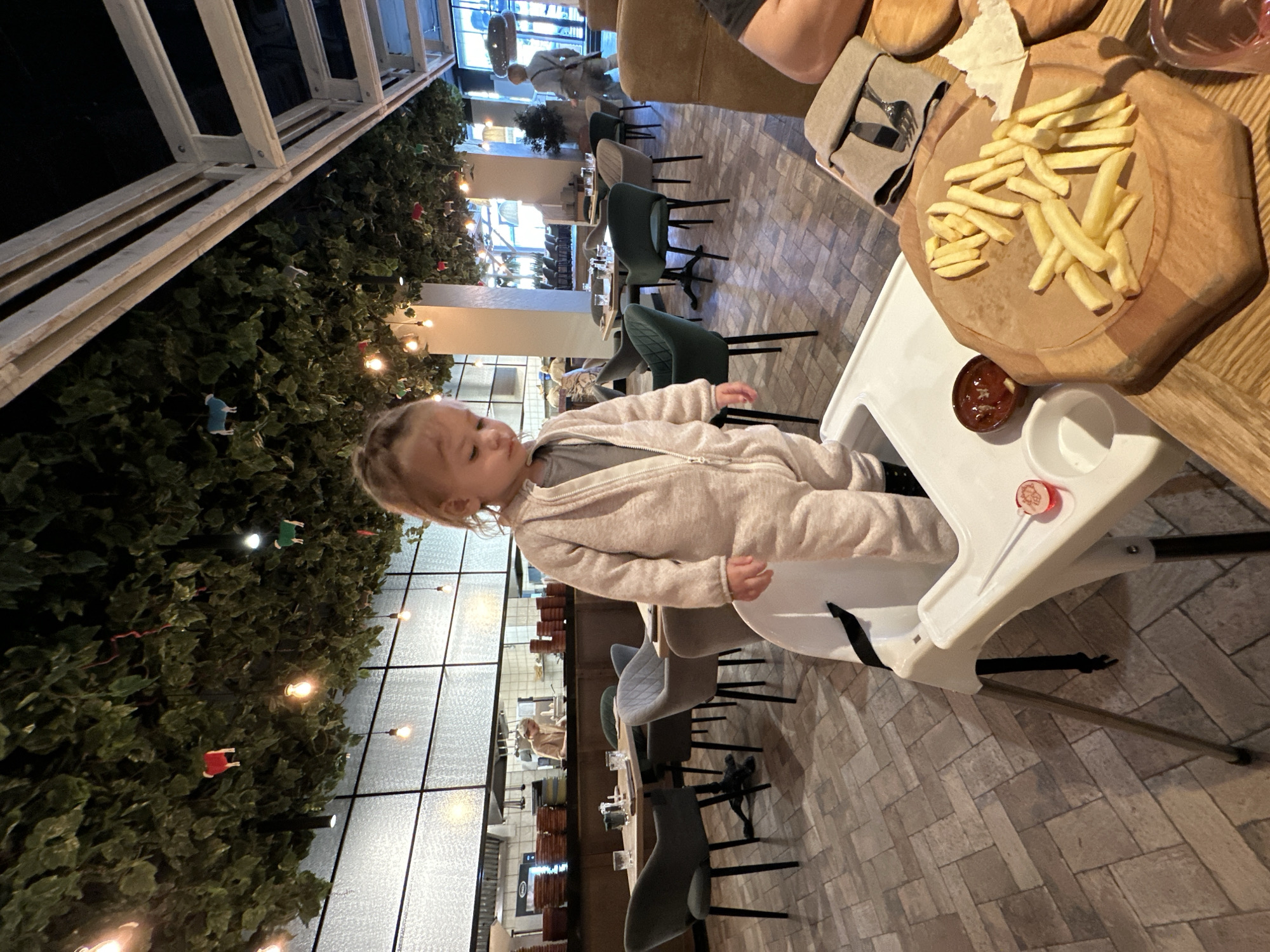 A child wearing a light-colored jumpsuit sits in a white highchair at a restaurant. A wooden cutting board holding french fries is on the table beside the highchair. The restaurant is likely located in Izhevsk, Russia. The background includes a wall covered with greenery and string lights, along with tables and chairs. Other people are visible further away.