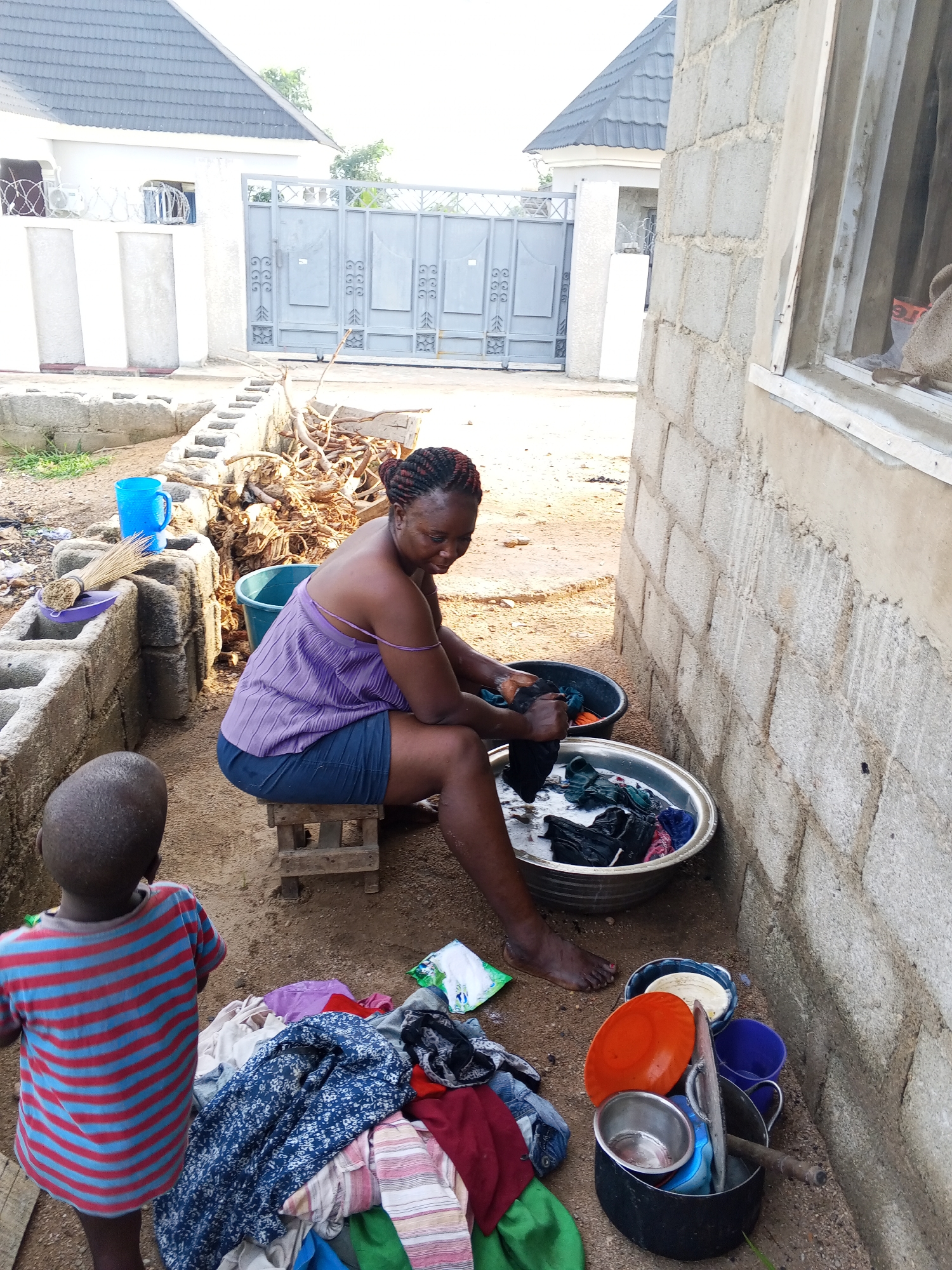 In an outdoor setting in Adonkolo, Nigeria, a woman is engaged in hand-washing clothes while a young child stands observing. The woman, with braided hair and wearing a purple spaghetti strap top and dark shorts, is seated on a small wooden stool. She is actively scrubbing clothes in a large silver basin filled with soapy water, with another basin containing dark clothes beside it. A substantial pile of mixed garments lies on the dirt ground in front of her.
To her immediate left, a low wall constructed from concrete blocks supports a blue plastic mug and a traditional broom. Beyond this wall, a large heap of dried branches or firewood is visible. The background features a tall, grey metal gate with decorative elements, leading to a property with white-painted walls and buildings topped with dark grey tiled roofs.
Near the woman's bare feet, a black pot holds an assortment of household items, including an orange plate, a small metal bowl, and a blue cup, suggesting other domestic tasks are being managed concurrently. The immediate ground is unpaved dirt. The child, viewed from the back, wears a red and blue striped t-shirt. The scene is brightly lit, indicating daytime and clear weather. The overall environment, with its concrete block construction and unpaved areas, is characteristic of a residential yard in a developing region. No prominent readable text is visible in the image, aside from a small, illegible label on a packet within the pile of clothes.