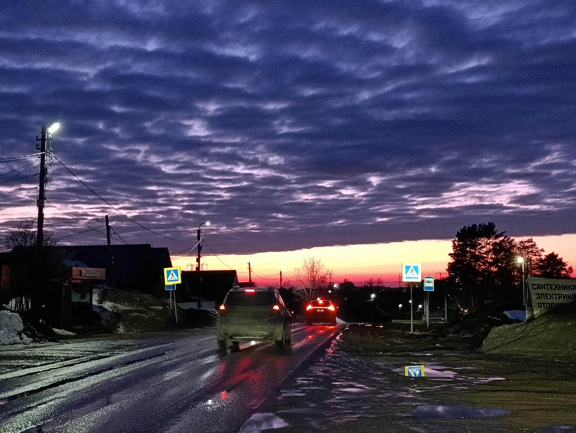 The image captures an exterior road scene at twilight in the city of Chaikovsky, Russia. The sky overhead features dark blue and purple altocumulus clouds, transitioning to a strip of pink and orange at the horizon, indicative of either sunrise or sunset. The asphalt road is wet and reflective, showing puddles and tire tracks.
Two dark-colored SUVs are visible from the rear, traveling away from the camera. The foreground SUV has illuminated red taillights reflecting on the wet road surface. A second SUV is further down the road, also displaying red taillights.
On the left side of the road, a utility pole with an active streetlight is present. Behind it are indistinct, dark buildings and piles of melting snow and dirt. A blue square pedestrian crossing sign with a white triangle and walking figure is clearly visible on a pole on the left.
On the right side of the road, further into the distance, another identical pedestrian crossing sign is visible, alongside a blue square sign with a white bus icon. A large, dark sign on the far right displays Cyrillic text: "САНТЕХНИКА" (Plumbing), "ЭЛЕКТРИКА" (Electrics), and "ОТОПЛЕНИЕ" (Heating), indicating a commercial enterprise. Patches of snow and dirt are present on the road shoulders. In the background, bare trees and some evergreen trees are silhouetted against the colorful sky, with additional illuminated streetlights visible further down the road. A yellow rectangular sign with a black border and an obscured symbol is on the ground near the right road edge.