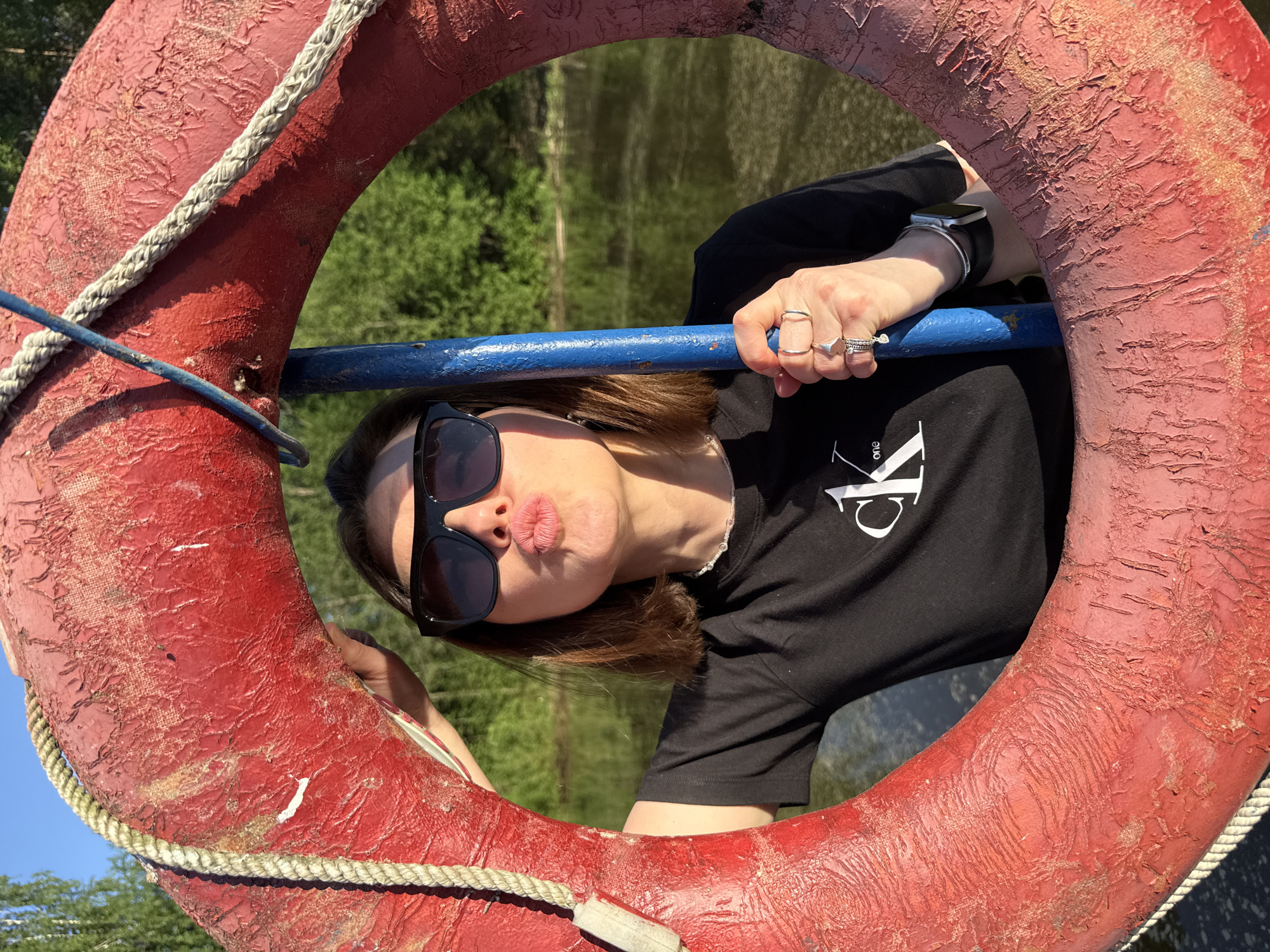A woman wearing sunglasses and a black t-shirt is posing inside a red life preserver. She is puckering her lips. She is holding onto a blue bar attached to the life preserver. The background shows a body of water and trees. The location is Chaikovsky, Russia.