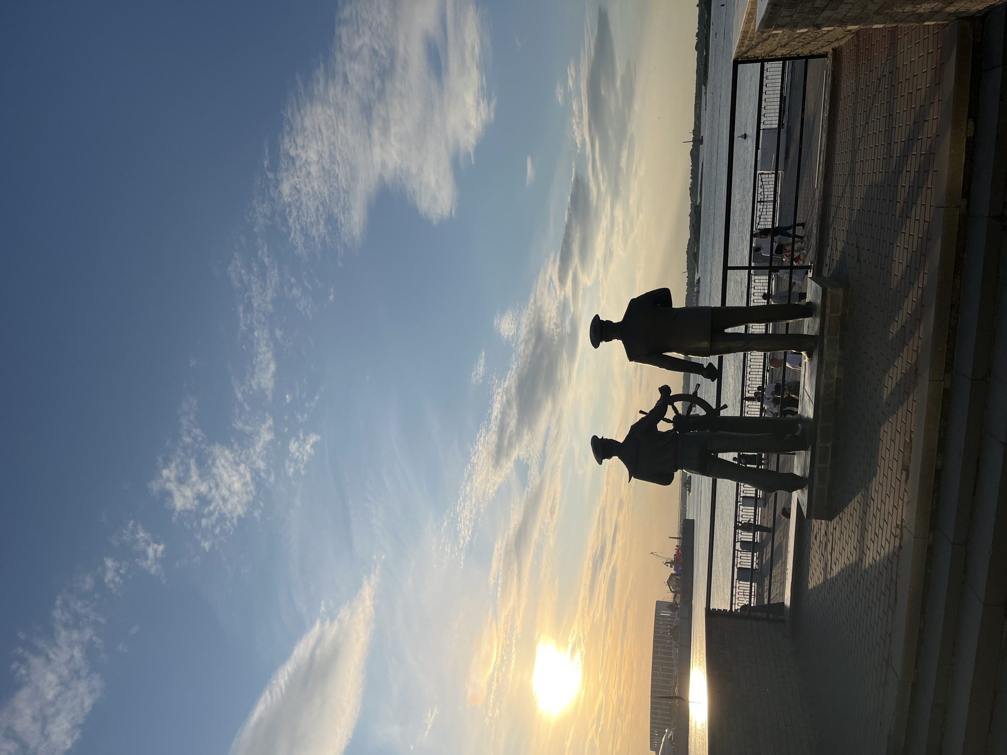 This is a vertical, outdoor, daytime photograph of a waterfront scene. The image captures two silhouetted statues of men in uniform, one holding a ship's wheel, standing on a brick pathway. They are positioned on a raised platform overlooking a body of water where several boats are visible, including a large ship and a crane in the distance. Beyond the water, a city skyline with buildings is discernable.
The sky is a clear blue with scattered white clouds, and the sun is low on the horizon, casting a warm golden glow that suggests either sunrise or sunset. The light creates long shadows on the pathway and illuminates the water. In the background, beyond a black metal railing, a few people can be seen walking and sitting, enjoying the waterfront. The overall atmosphere is peaceful and scenic, with the statues providing a historical or commemorative element to the modern urban landscape. There is no visible text in the image.