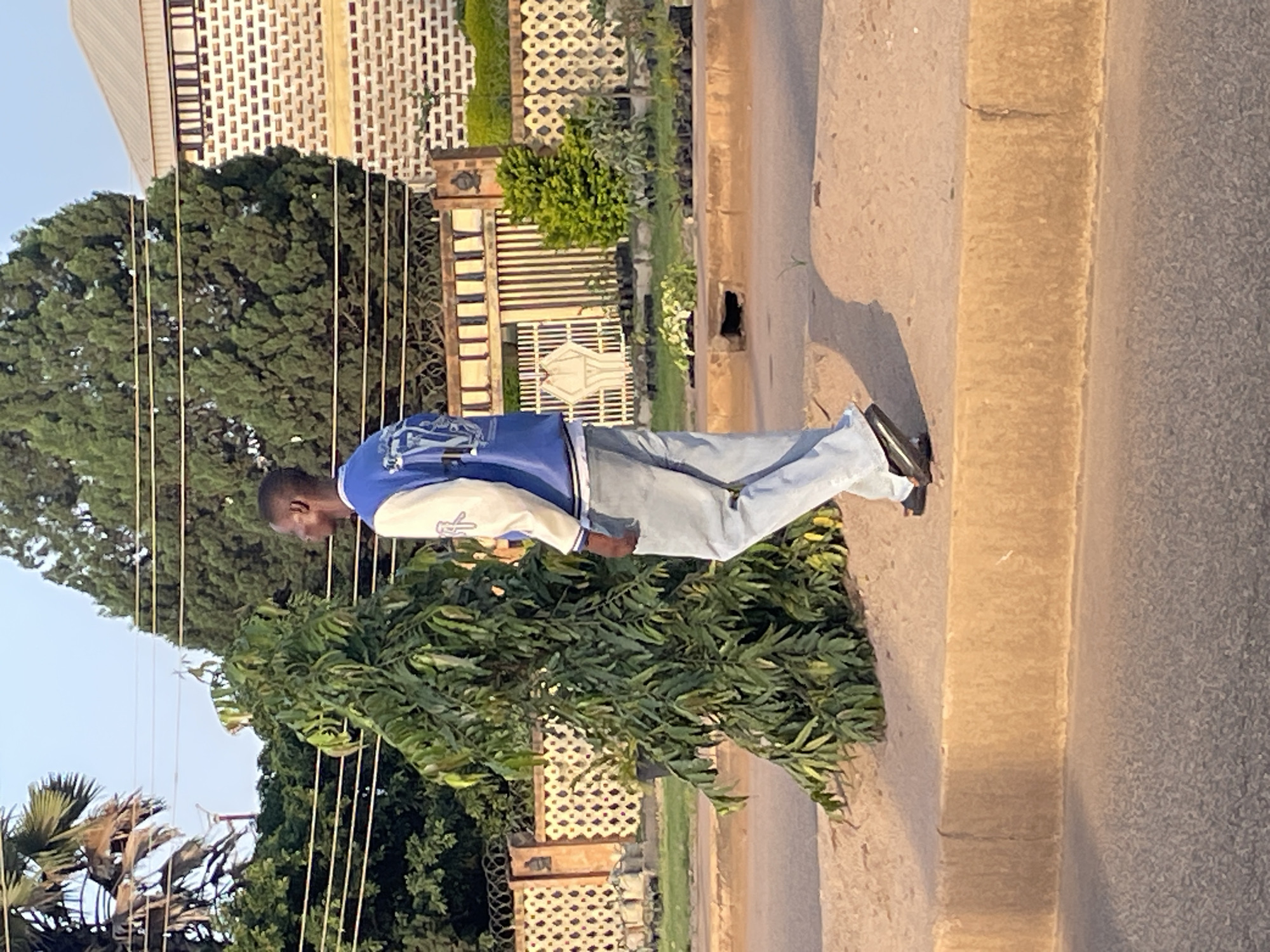 A young Black man walks along a dirt and asphalt road in Jos, Nigeria, beside a decorative concrete curb and a small tree. He is wearing a blue and white letterman jacket, light blue jeans, and black sandals. The background features a white brick building with a patterned facade, a metal fence, and lush green trees under a bright sky. The lighting suggests it is either early morning or late afternoon.