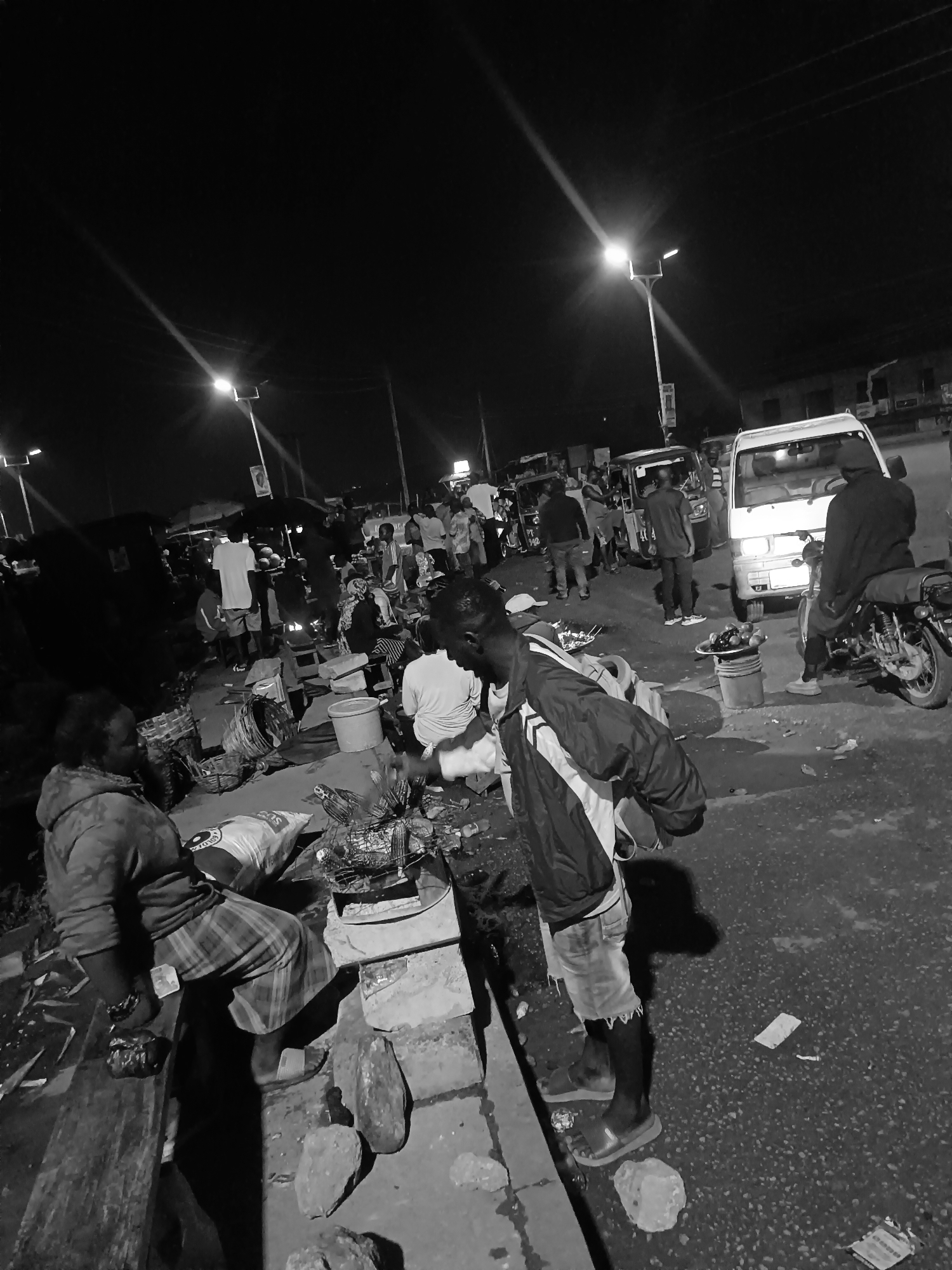The image depicts a nighttime scene at what appears to be an outdoor market in Bukuru, Nigeria. The area is illuminated by streetlights, though the overall lighting is dim. The foreground features a woman sitting on a low bench with what appears to be merchandise laid out for sale. Nearby, a man is standing, seemingly inspecting or purchasing goods from another vendor behind a low wall. The market area is crowded with people milling about, and various vehicles, including a white van and several motorcycle rickshaws (tuk-tuks), are parked on the road. Debris litters the street. The general atmosphere suggests a bustling informal market in a densely populated area.