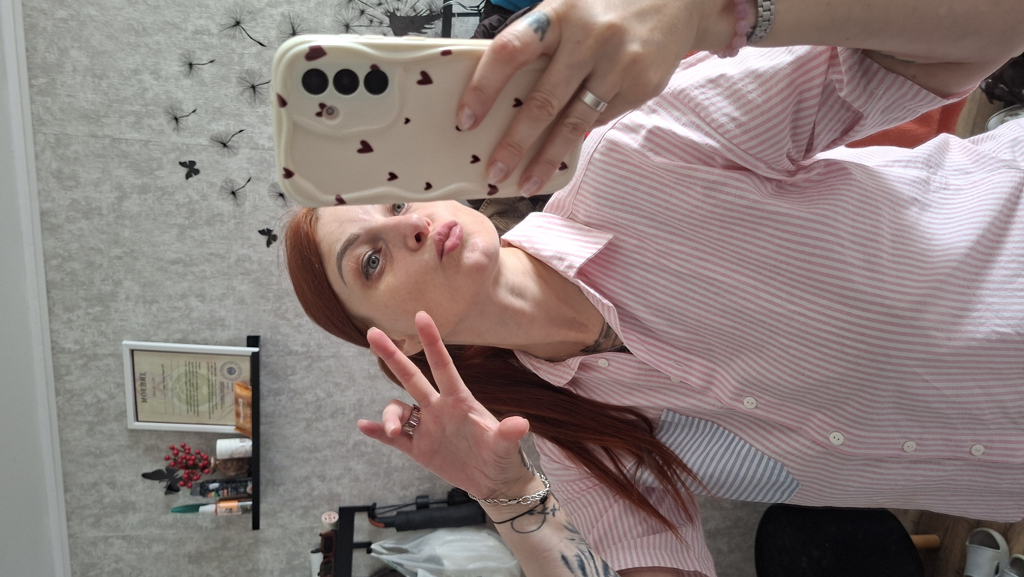 A medium shot captures a woman with red hair taking a mirror selfie in a room. She is wearing a pink and white striped button-down shirt and has a phone with a heart-patterned case in her right hand. Her left hand is held up, fingers spread in a peace sign, and she has rings on both hands. A bracelet is visible on her left wrist. In the background, a shelf holds a framed certificate, a small vase with red berries, and other items. The wall behind her is decorated with a pattern of dandelions and butterflies. The scene appears to be indoors, likely a home environment in Myadzyel, Belarus, suggested by the provided context.
