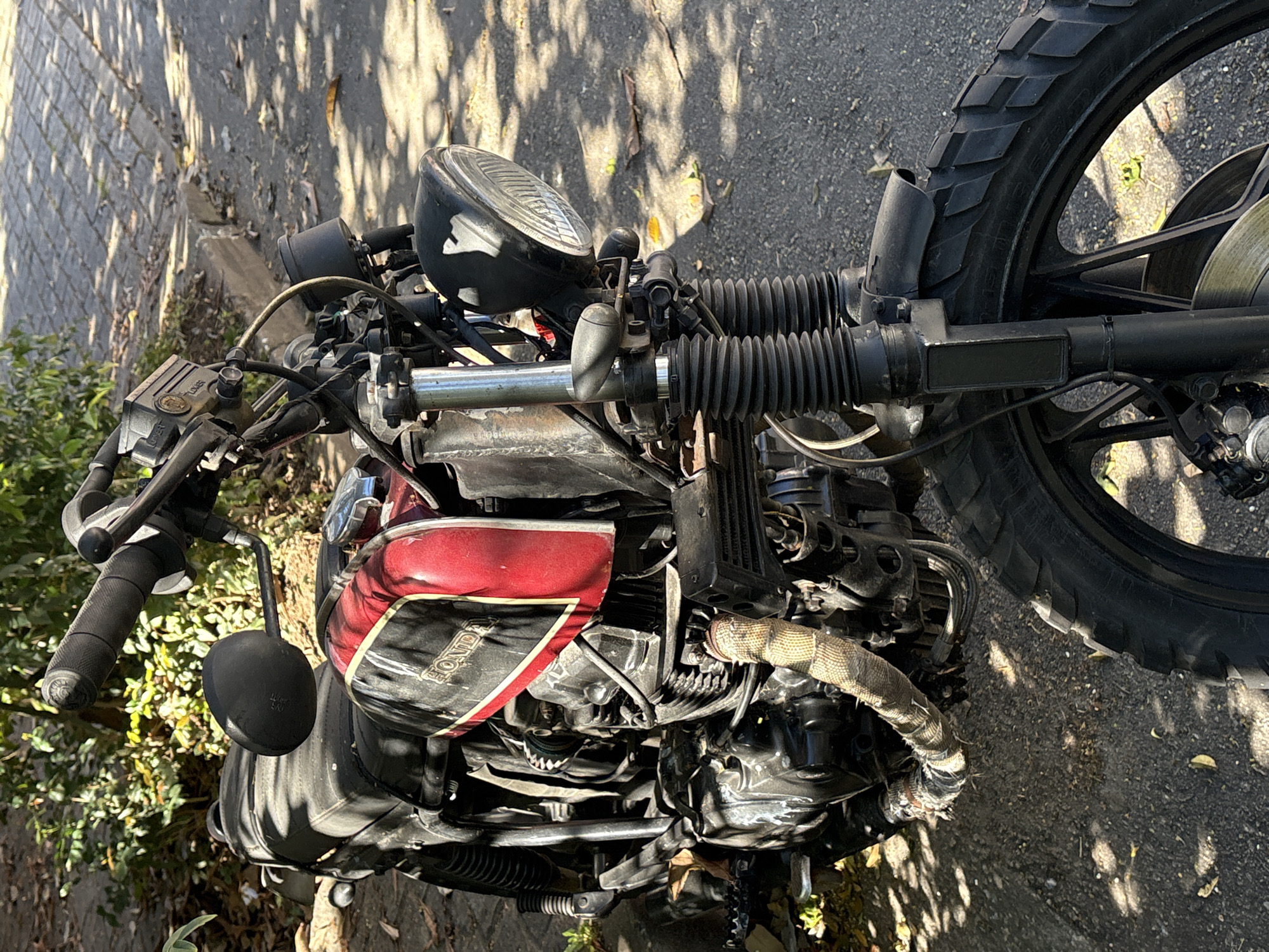 The image shows a close-up, eye-level view of a vintage red and black Honda motorcycle parked outdoors on a paved surface with scattered leaves and sunlight dappling through overhead foliage. The motorcycle is positioned diagonally, with its front wheel and engine area most prominent. Details visible include the handlebars, a black rearview mirror, the red fuel tank with a yellow and black stripe and "HONDA" lettering, the engine block, exhaust pipes wrapped in heat-resistant material, front suspension forks, a black headlight, and a portion of the knobby front tire. The background consists of greenery and a cobblestone-like pathway with shadows. The scene suggests a calm, daytime setting, likely a street or sidewalk in an urban area. The presence of leaves and dappled sunlight indicates it is likely autumn or a season with deciduous trees. No people are visible in the image. The location is identified as the city of São Paulo, Brazil.