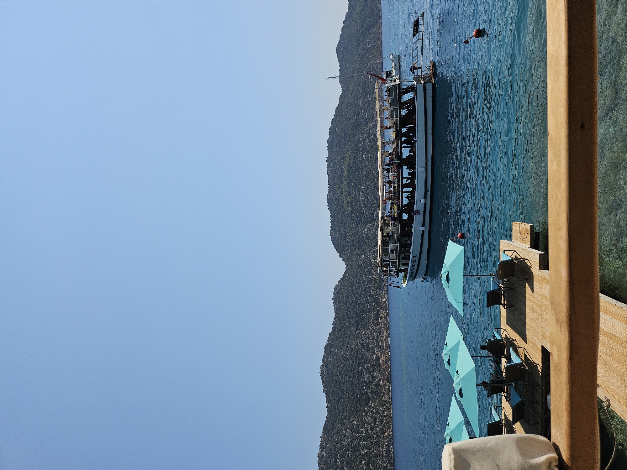 An eye-level, vertical shot captures a bright, sunny day at a coastal location. In the foreground, a wooden dock with a light brown railing is visible, partially obscuring a section of the dock with turquoise umbrellas and empty lounge chairs. To the right of the dock, the clear blue water reveals submerged rocks and a light brown rope.
Further out in the water, a large white tour boat, identified by the name "NESKO 2" on its side, is filled with passengers. The boat has multiple decks and a canvas covering. To the right of the tour boat, a smaller white sailboat is anchored. In the background, lush green hills rise from the water's edge under a clear, pale blue sky. Two red buoys are floating in the water.
