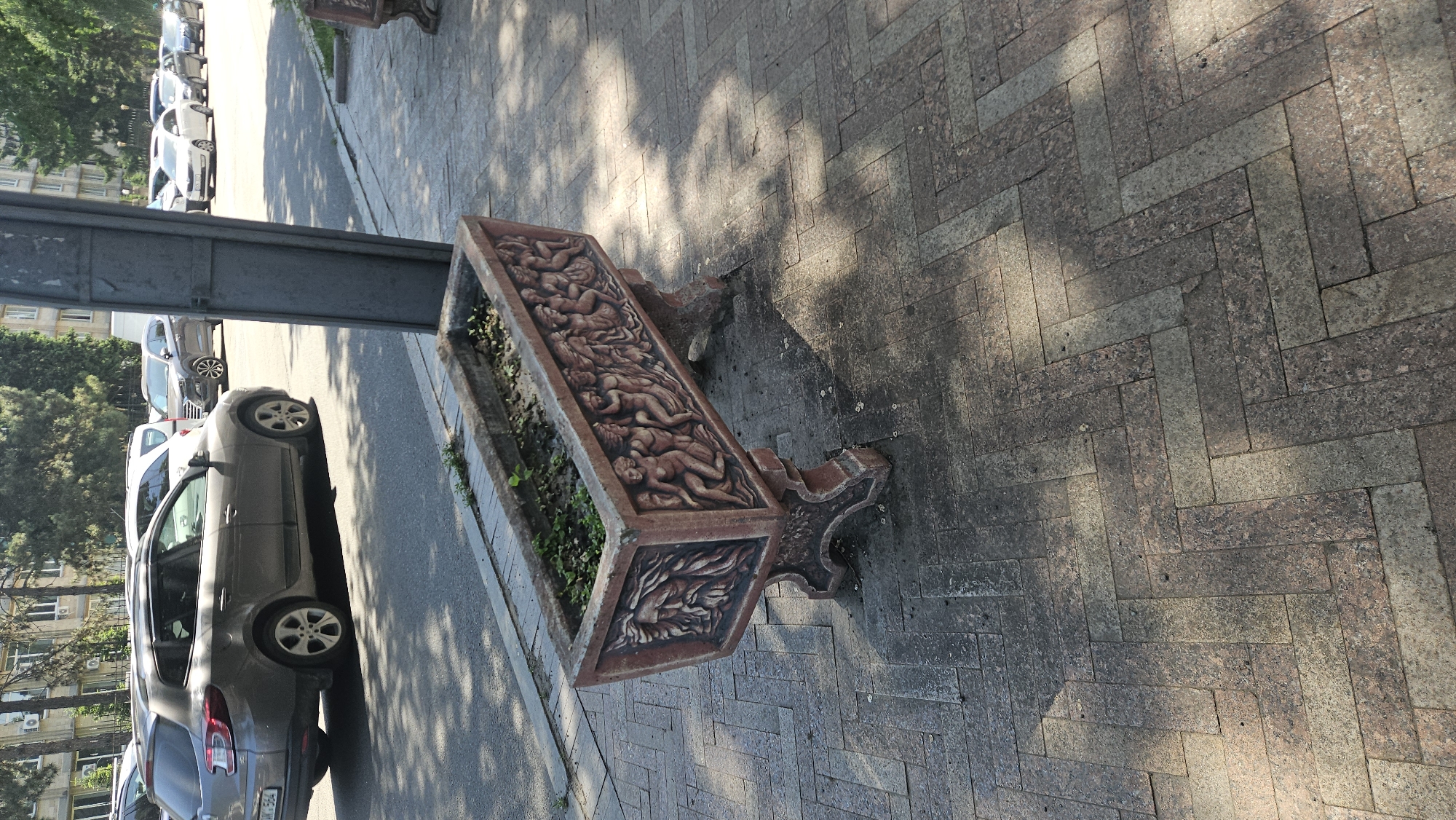 A photograph shows a street scene in Chișinău, Moldova. A large, ornate terracotta flower planter sits on a herringbone-patterned brick sidewalk. The planter is decorated with bas-relief figures and contains sparse greenery. A grey metal pole is near the planter, suggesting a possible bus stop. Cars are parked along the adjacent street, and buildings with trees are across the street. The time of day is likely mid-morning or mid-afternoon, as suggested by the light and shadows.