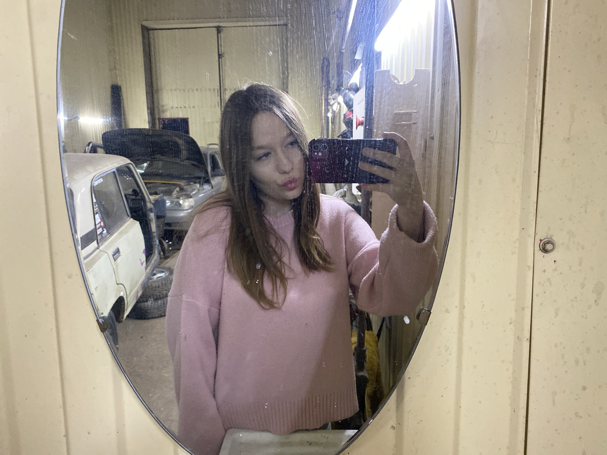 A young woman with light brown hair is taking a selfie in a dirty, oval mirror. She is wearing a pink sweater and has a slightly puckered expression. In the mirror's reflection, a garage or workshop setting is visible. There are several vehicles, one of which is a light-colored car with its door open, and another with its hood raised. Discarded tires are stacked in the background. Based on the metadata, this image was taken in the city of Chaikovsky, Russia. The mirror itself is mounted on a grimy, light-colored, possibly metal wall. The scene appears cluttered and industrial.