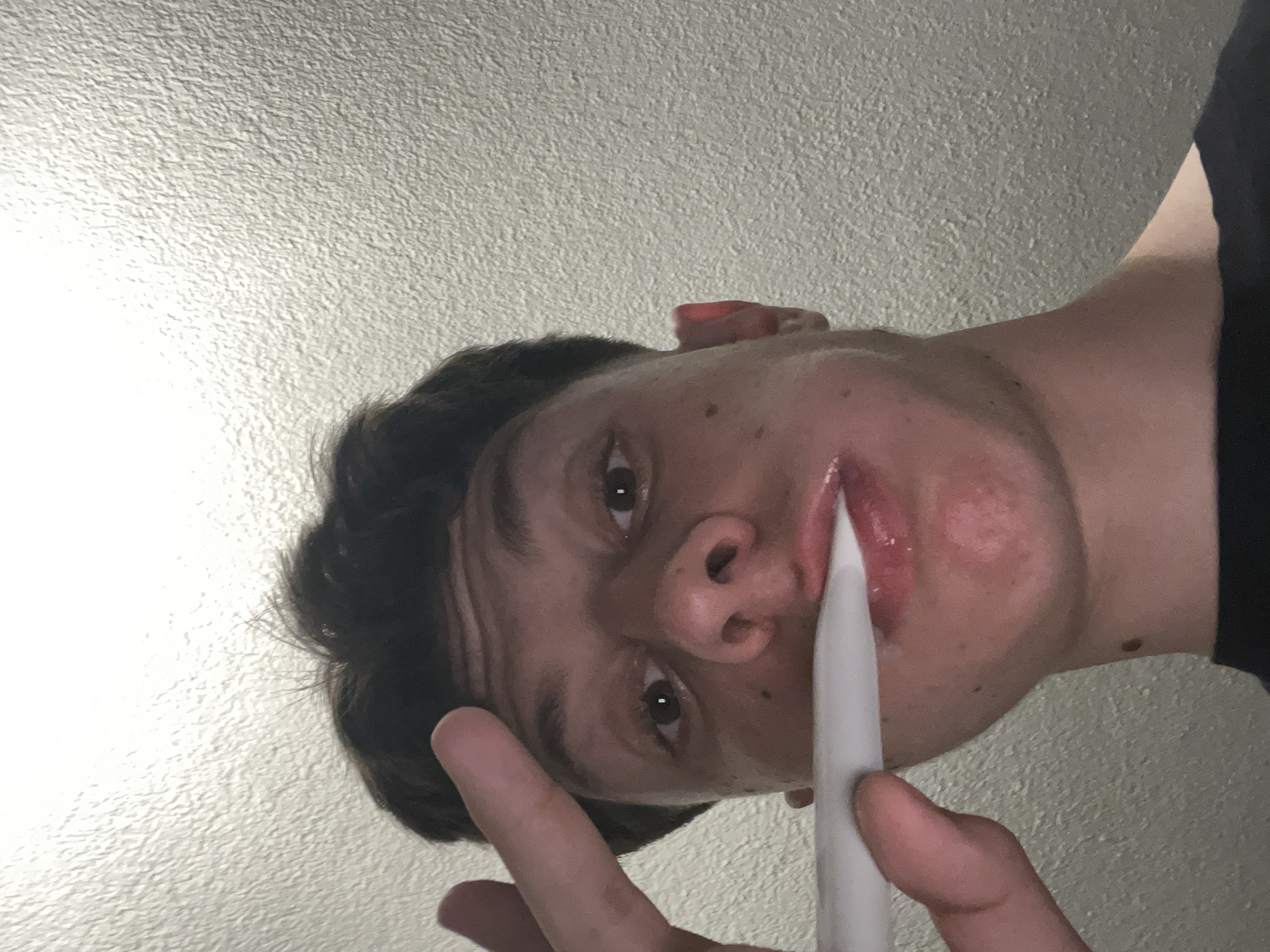 The image is a close-up, low-angle, indoor shot of a young male. He has dark, short, curly hair and brown eyes. His skin is light-toned with some minor blemishes. He is holding a white object, possibly a stylus or pen, up to his mouth, with his index finger and thumb pinching it. His lips are slightly parted and appear glossy. The background is a textured, light-colored wall. The image does not provide any information that would allow for the determination of its location in Zlatograd, Bulgaria, or any other specific city.