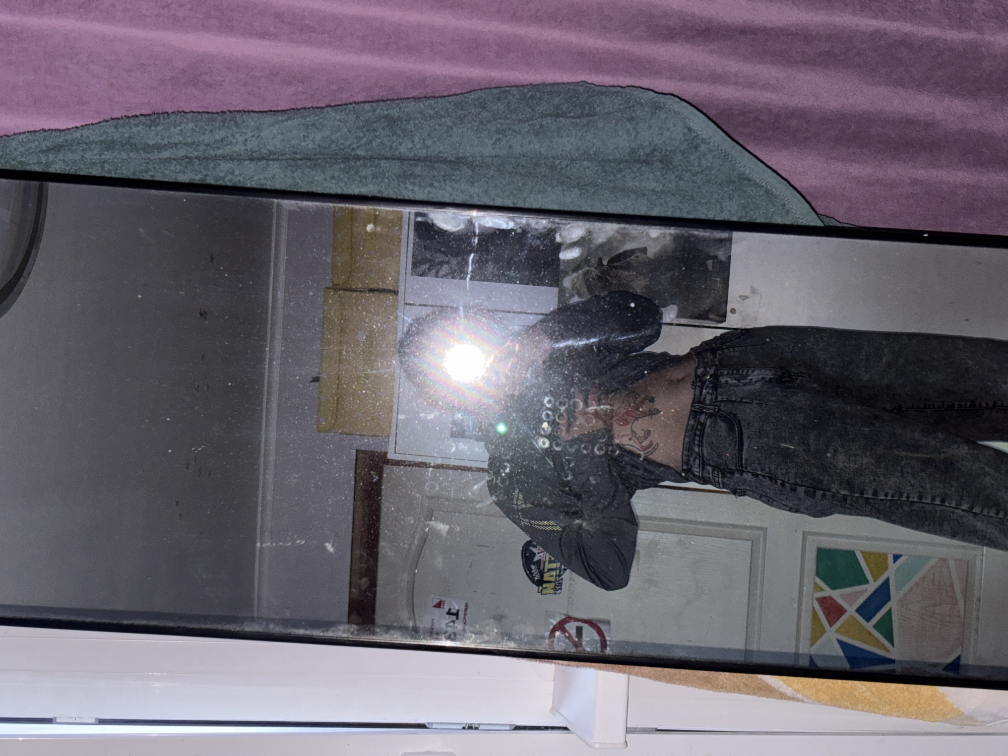 A full-length mirror selfie captured in a room in Прикамский, Russia. The black-framed mirror is notably dusty, showing numerous small white specks across its surface. A bright camera flash originating from the individual's device obscures their head and upper chest, casting a strong glare. The individual is wearing a dark, long-sleeved top that appears sheer or mesh over the abdomen, revealing multiple tattoos. A prominent red-brown tattoo depicting an animal (possibly a dog or wolf) in profile is visible on the right side of the individual's ribcage. They are also wearing dark, high-waisted acid-wash jeans.
Reflected in the mirror is a white paneled door featuring an inverted word at the top, a red circular "No Smoking" sign, and a "NOON" sticker. A colorful abstract geometric artwork is affixed to the lower panel of the door. Above the door, several yellowish-brown boxes are stacked. To the left of the mirror, a white door frame or window frame with a small white shelf is visible. To the right of the mirror, a green textured towel hangs against a pink or purple corrugated curtain or wall.