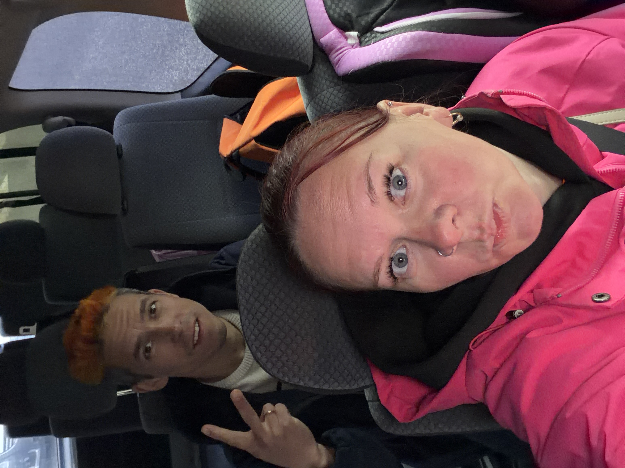 This media file captures a candid photo taken inside a vehicle, appearing to be a car or minivan, with the image rotated 90 degrees counter-clockwise.
In the foreground, a woman with fair skin, striking blue eyes, and reddish-brown hair is featured, leaning against a gray fabric headrest. She is wearing a vibrant pink hooded jacket over a black top, and a silver septum nose piercing is visible. Her expression is somewhat pouting or playful.
Behind her, positioned further back in the vehicle, is a man with short, bright orange hair. He has fair skin and is wearing a light-colored sweater beneath a dark jacket. He looks towards the camera with a slight smile and makes a peace sign gesture with his left hand.
The interior of the vehicle includes gray patterned fabric car seats and headrests. An orange bag is visible behind the woman's head, and a purple and black bag can be seen in the upper right portion of the frame. Through the window, a blurred, speckled background suggests either motion or precipitation on the glass. The scene appears relaxed and informal, possibly capturing a moment during a journey in Izhevsk, Russia. No visible text is present in the image.