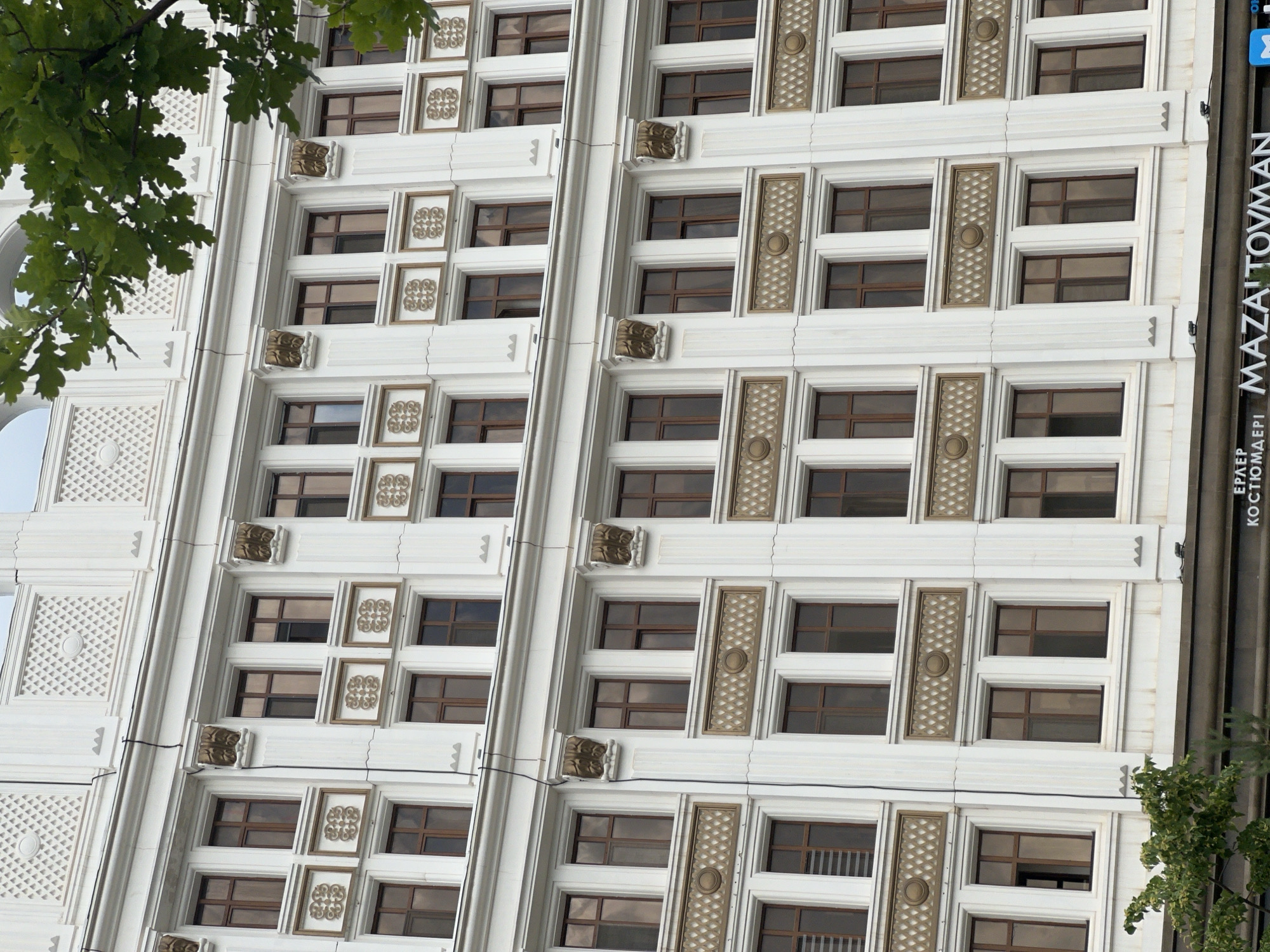 The image shows a section of a white multi-story building with ornate detailing. The building has a repetitive pattern of windows, with rows of rectangular windows divided by decorative columns and horizontal bands. The windows themselves have brown frames and appear to be tinted. There are also decorative elements above some of the windows, including what look like golden carvings. At the very bottom of the image, a dark band runs across the building with text visible, indicating it is a commercial establishment. Green leaves from a tree partially obscure the top right portion of the building. The architectural style suggests it is located in a city known for its grand and modern structures, such as Astana, Kazakhstan. The scene is static, with no people or actions depicted, focusing entirely on the architecture of the building.