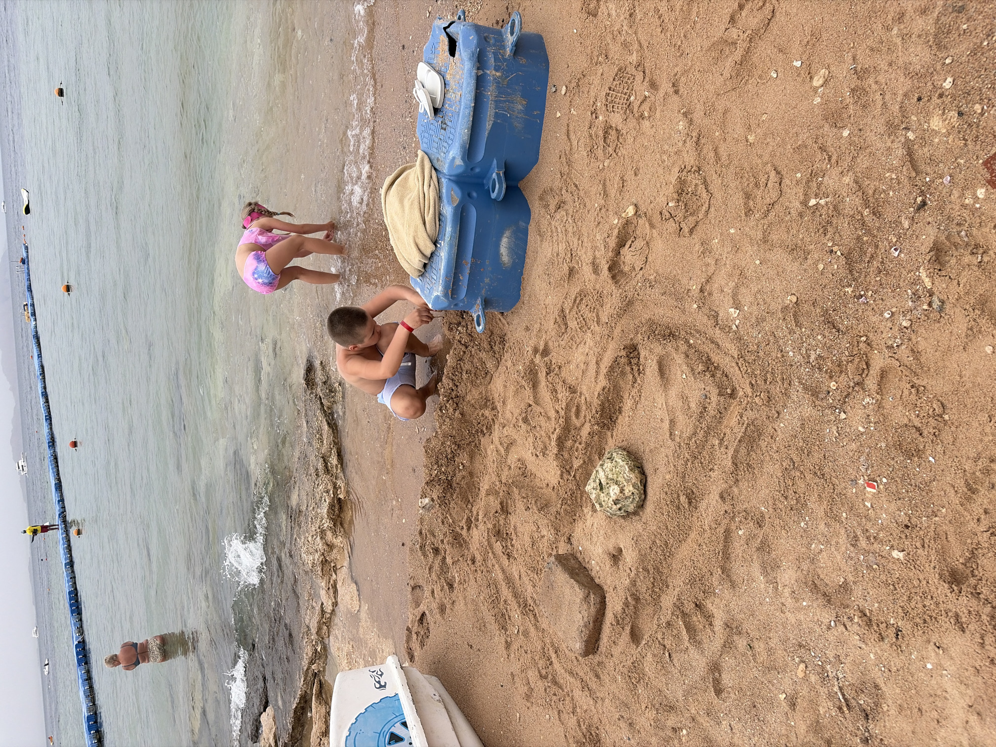 The image depicts a beach scene, possibly in Sharm El Sheikh, Egypt, based on the coastal environment. Two children are near the water's edge: one girl is leaning over, possibly touching the water, while a boy is kneeling near a blue plastic structure on the sand. A beige towel is draped over the structure. On the sand, there's a rock and a short, rectangular sand structure. In the water, a person is standing near a barrier with buoys. A lifeguard is visible in the background. The beach has brown sand with various small objects scattered on it.