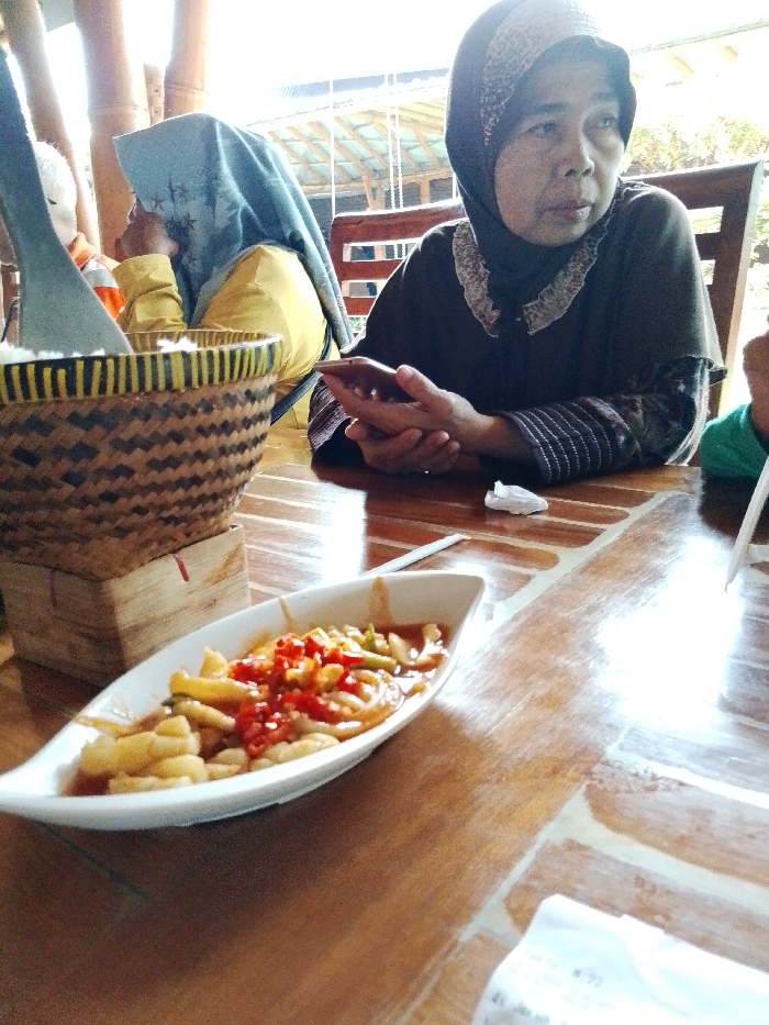 The scene depicts an interior dining area of a restaurant in Pabelan, Indonesia. A glossy, light-brown wooden table occupies the foreground, featuring visible grain and lighter-toned inlaid patterns. On the table, a white, boat-shaped ceramic dish contains a reddish-orange saucy food item with numerous light-colored pieces, red chili slices, and green vegetable fragments. To the left of the dish, a crumpled white paper napkin and a thin white straw or stick are present. Further left, a large, conical woven bamboo basket, featuring dark and light yellow striped banding and a dark woven pattern, holds a light-colored substance and a gray serving utensil. This basket rests on a light-colored square wooden base.
An adult female is seated at the table on the right side of the frame. She wears a dark hijab with a patterned band, a dark long-sleeved shirt with a light-colored, possibly embroidered, collar, and patterned cuffs. Her hands are clasped, holding a smartphone, and her gaze is directed towards her right. To her left, partially obscured by the basket, a second adult female is visible, wearing a light blue hijab and a yellow long-sleeved top, facing towards the left. A segment of a head is partially visible behind the basket on the far left. On the far right edge, a portion of an arm wearing a green shirt is present. The background reveals additional wooden seating, including chair backs with horizontal slats, and structural elements made of bamboo, with bright natural daylight entering the space from an open area.