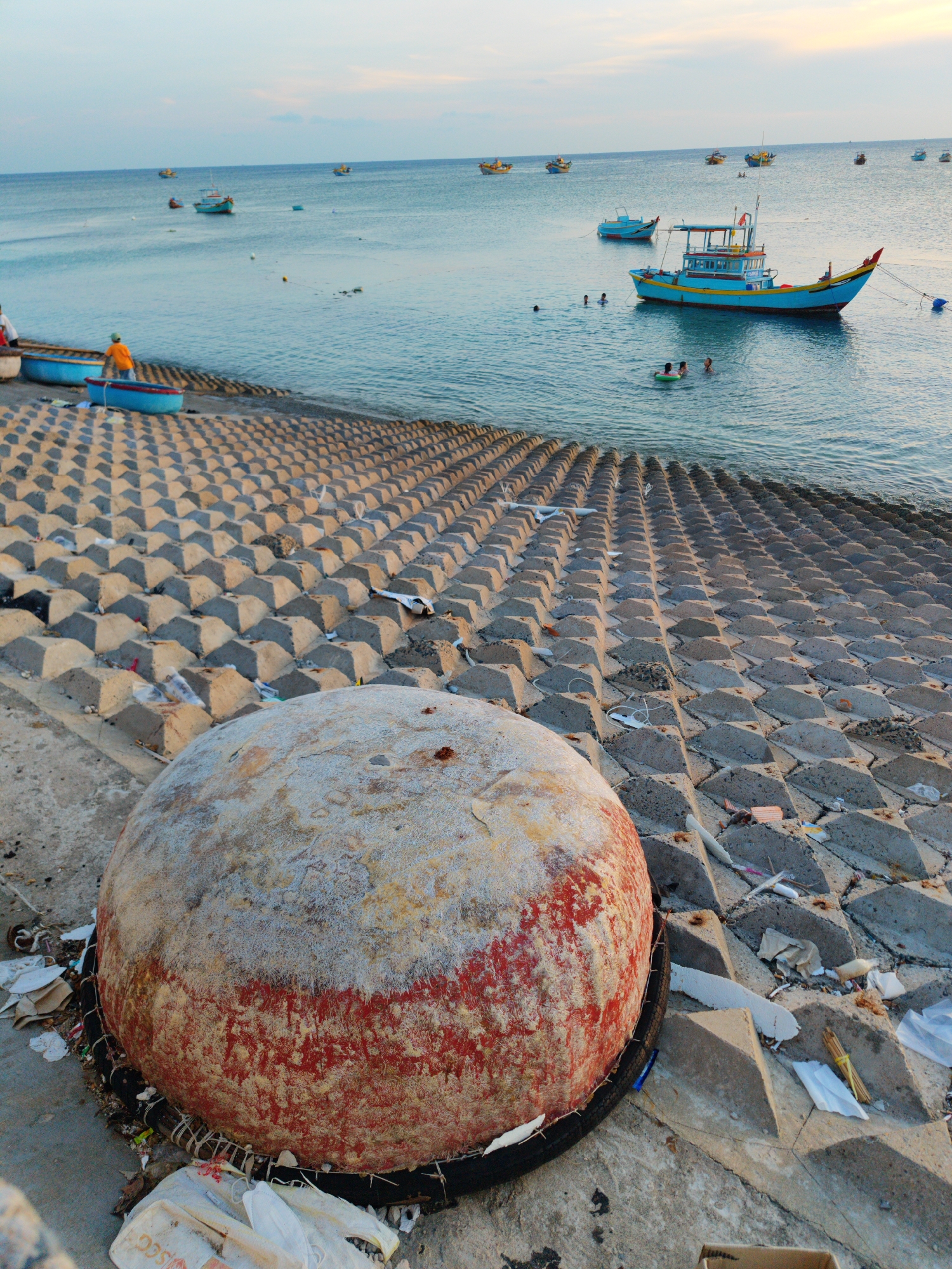The image shows a coastal scene in Phú Quý Special Administrative Region, Vietnam. In the foreground is a large, spherical buoy with a weathered white and red exterior, resting on a concrete surface encircled by a black tire. Litter and debris surround the base of the buoy. Behind the buoy is a concrete embankment constructed of interlocking pyramidal blocks leading down to the sea. The embankment is also littered with trash. The sea is calm with several boats anchored in the distance. In the water, some people are swimming. One small blue boat with an occupant in an orange shirt is near the shore. Another larger blue boat with yellow and red trim is closer to the swimmers. The sky is light blue with hints of pink, suggesting either sunrise or sunset.