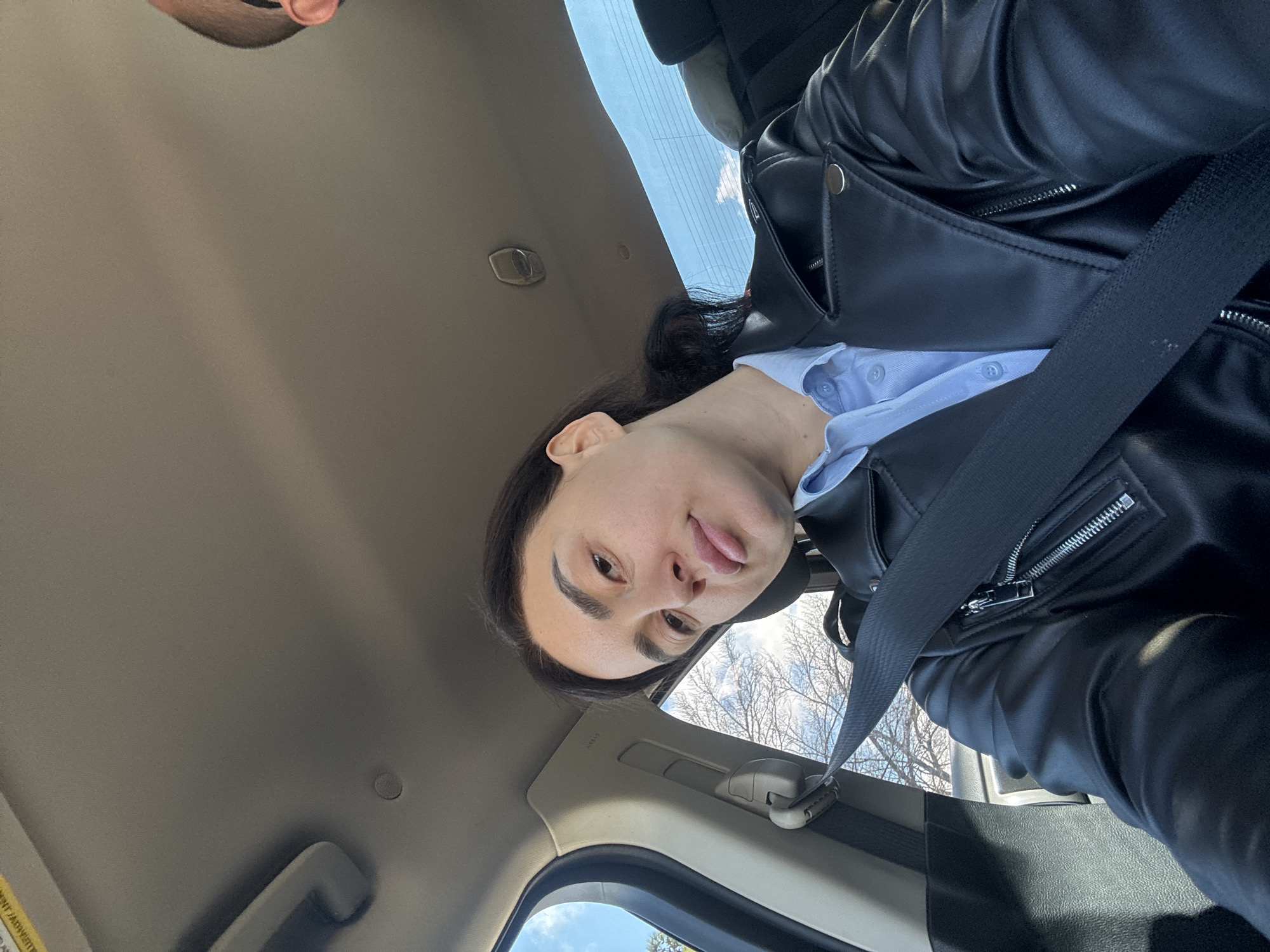 A person with dark hair, wearing a blue button-down shirt and a black leather jacket, is wearing a seatbelt in a car. The photo is taken from a low angle inside the vehicle, showing part of the car's interior, including the roof and door frame. Outside the window, bare tree branches and a glimpse of a cloudy sky can be seen. The location in Tbilisi, Georgia cannot be determined from the given visual information.