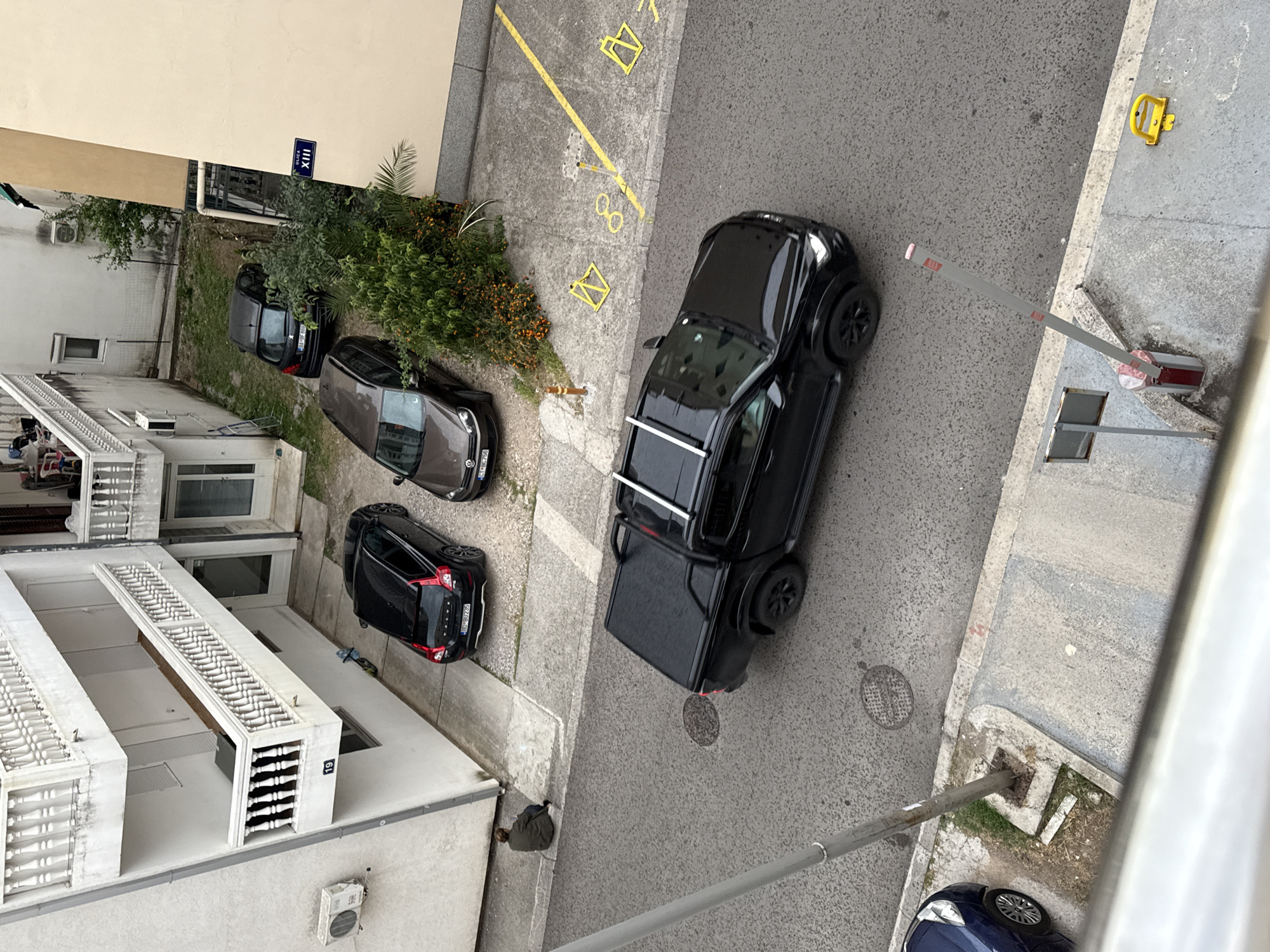 The image, taken from an elevated perspective, shows a street scene in Budva, Montenegro. Along the left side of the frame, there are multi-story buildings with white balconies and traditional window designs. Three cars are parked alongside the buildings: a dark grey car, a small black hatchback, and another black car. A street sign labeled "XIII" is visible on the adjacent building.
Parallel to these buildings, a paved street runs from the upper-left to lower-right. A black pickup truck with a covered bed is parked at an angle to the road. Adjacent to the pickup truck on the right is a parking barricade, and further along the street, a yellow parking bollard is bolted to the sidewalk. The street surface has visible manhole covers.
To the right of the road, a narrower sidewalk runs alongside a taller building. A blue vehicle is visible in the bottom-right corner of the frame. The overall impression is of a quiet residential area with typical urban street features. A person is standing by a building near the bottom left of the image.