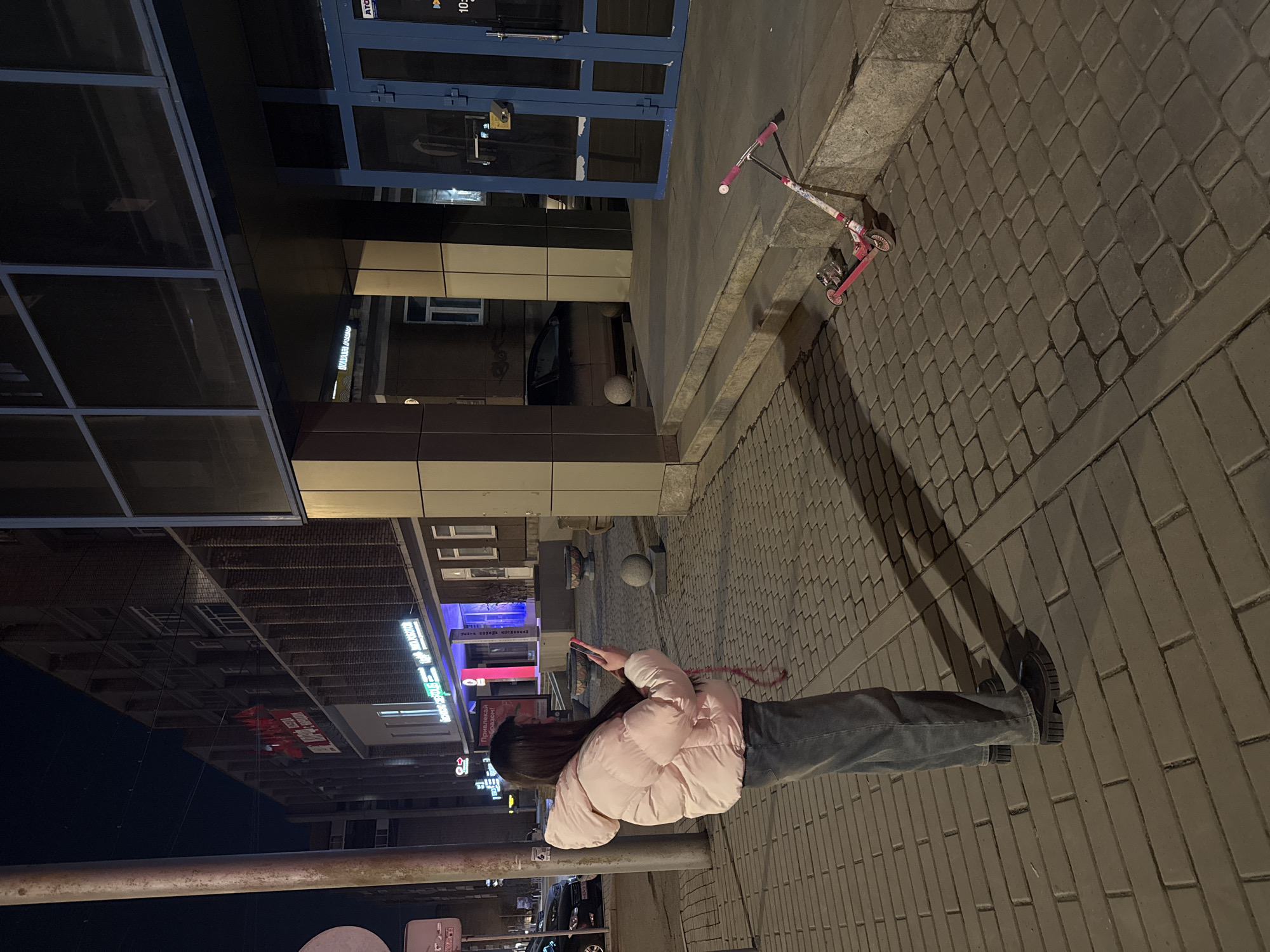 A medium, tilted shot taken at night in Krasnoyarsk, Russia. The composition features a woman wearing a light pink puffer jacket and jeans standing on a brick sidewalk. She is facing left, with her head near a pole, holding what appears to be a phone in her hands. Adjacent to her on the right, extending toward the top of the frame, the brick sidewalk continues. In the upper right corner is a child's scooter, in front of a building with blue-framed windows. The opposite side of the frame shows city buildings, signage in Russian, and cars, visible due to reflections in the building windows.