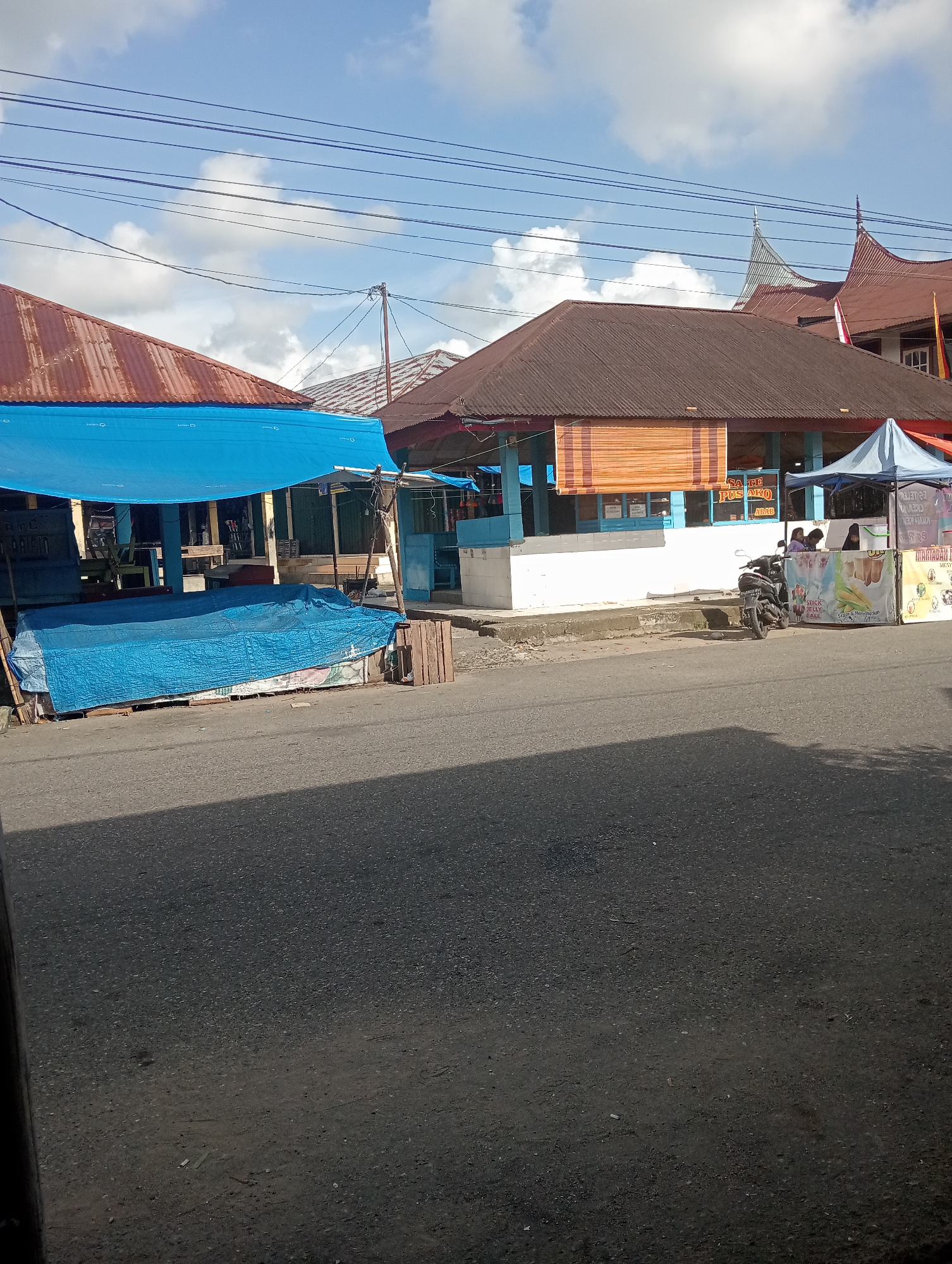 The image depicts a street scene in Pariaman, Indonesia. On the left, a market stall is covered by a large blue tarp, with another blue tarp covering goods on the ground. The stall has a corrugated metal roof. To the right, a building with a brown corrugated metal roof and blue trim is visible. This building appears to be a shop or eatery, with bamboo blinds and windows. A blue canopy tent is set up in front of this building. A motorcycle is parked near the building, and two women are seated behind a counter. Banners with text and images are displayed in front of the tent. The street in the foreground is paved asphalt, and the sky above is blue with scattered white clouds. Overhead power lines are strung across the sky. The overall scene suggests a casual, daytime market or commercial area.