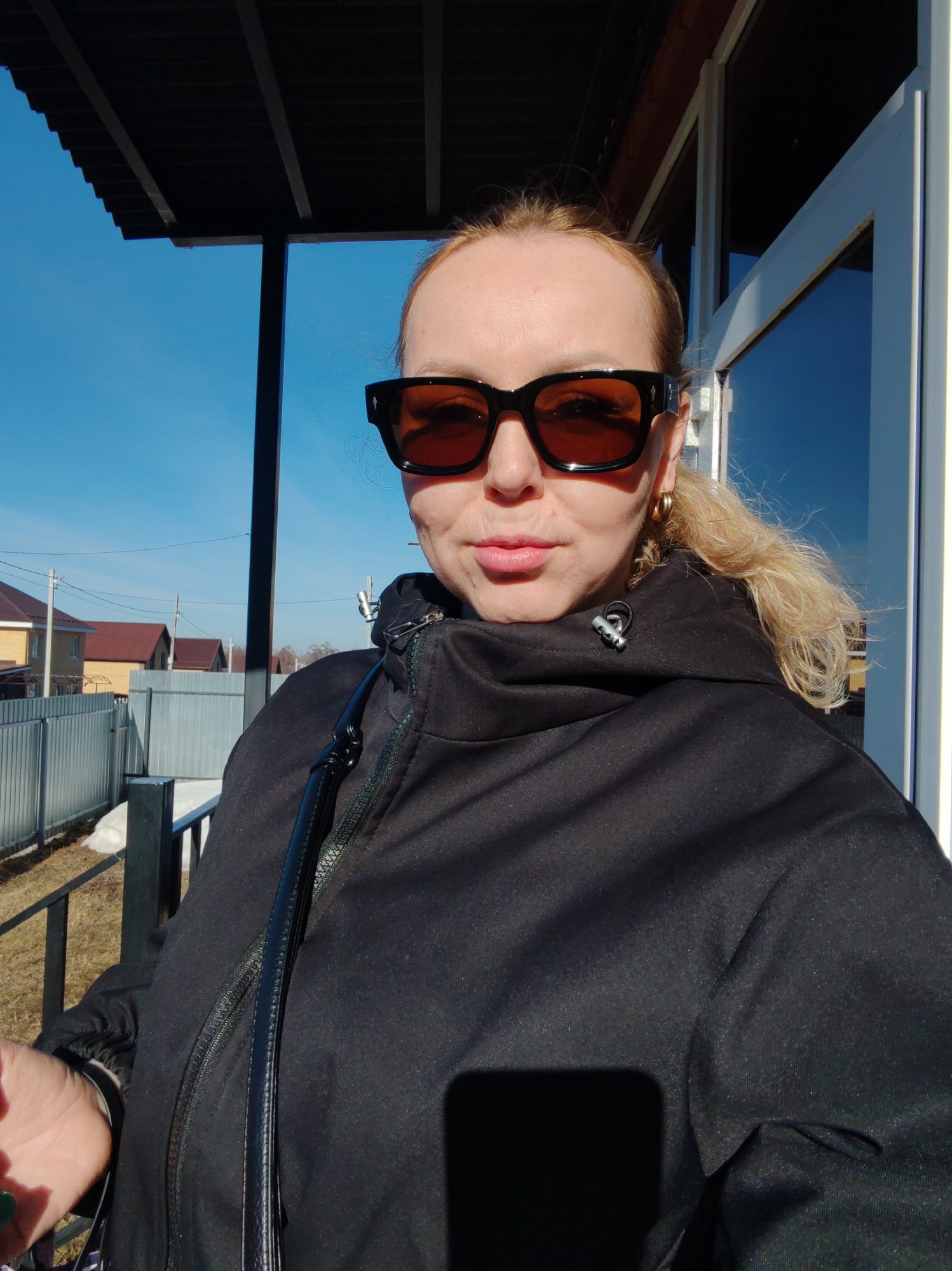 A blonde woman wearing sunglasses and a black hooded jacket is posing for a selfie outdoors in Chaikovsky, Russia. Behind her is a partially visible house with a white door frame and a mirrored window reflecting a clear blue sky. To the left, there's a view of neighboring houses with brown roofs and a grey fence. The ground appears to have patches of snow or ice. The woman is wearing gold earrings.