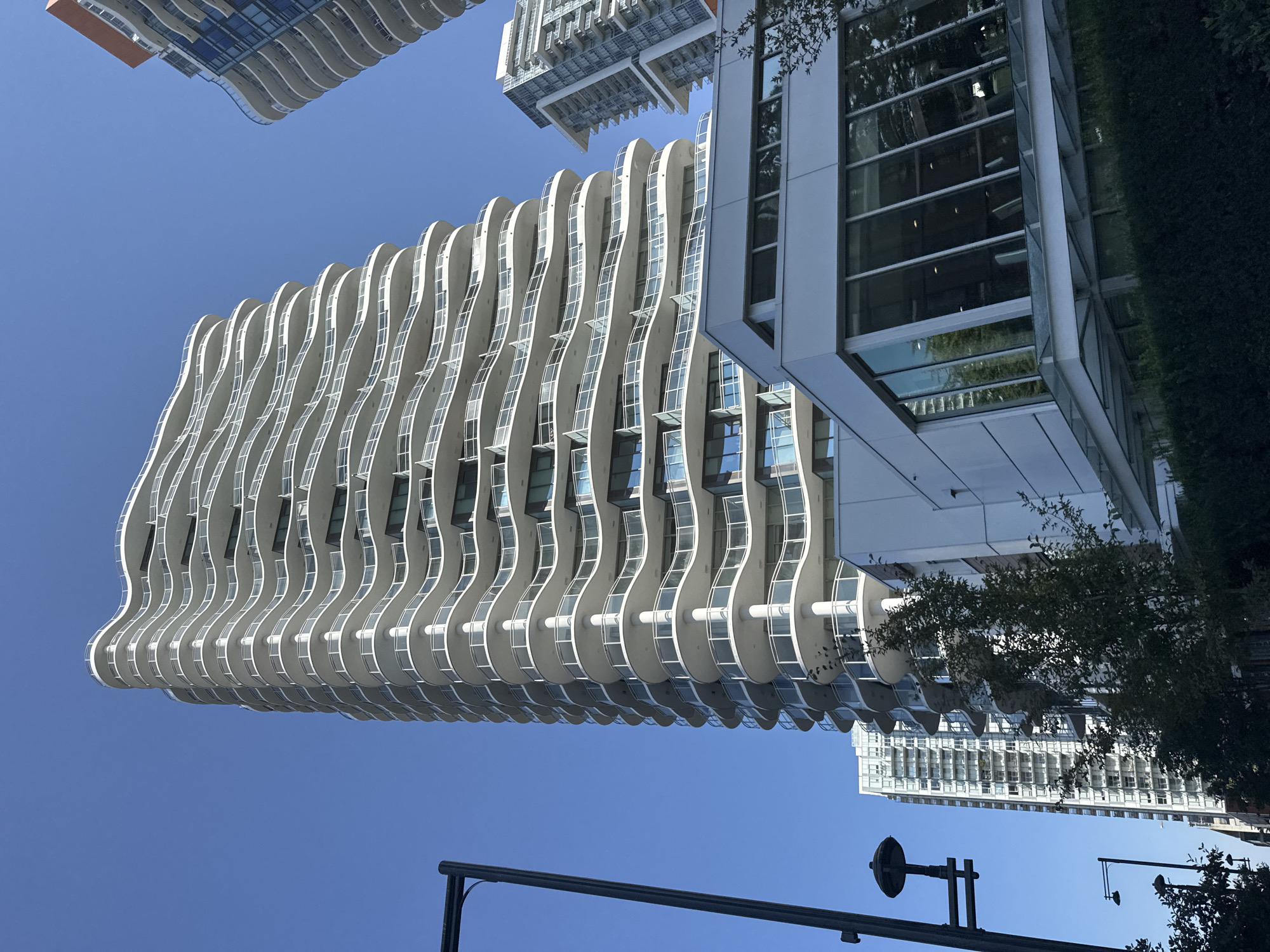 This is a low-angle, full shot of the Serenity, a residential building located in Surrey, Canada, on a clear, sunny day. The building features a distinctive, undulating facade with white balconies that create a wave-like pattern. The balconies are enclosed by clear glass railings. The building is tall and modern, with visible windows and doors on each floor. In the foreground, a contemporary glass-enclosed structure is partially visible, connected to a larger white building. Trees and landscaping are present in front of the lower structure. To the right and behind the main building, another modern high-rise building with a contrasting darker facade and balconies is partially visible. To the left of the main building, a dark metal lamppost with a decorative fixture is visible, and further in the distance, another lamppost. The sky is a clear, vibrant blue.